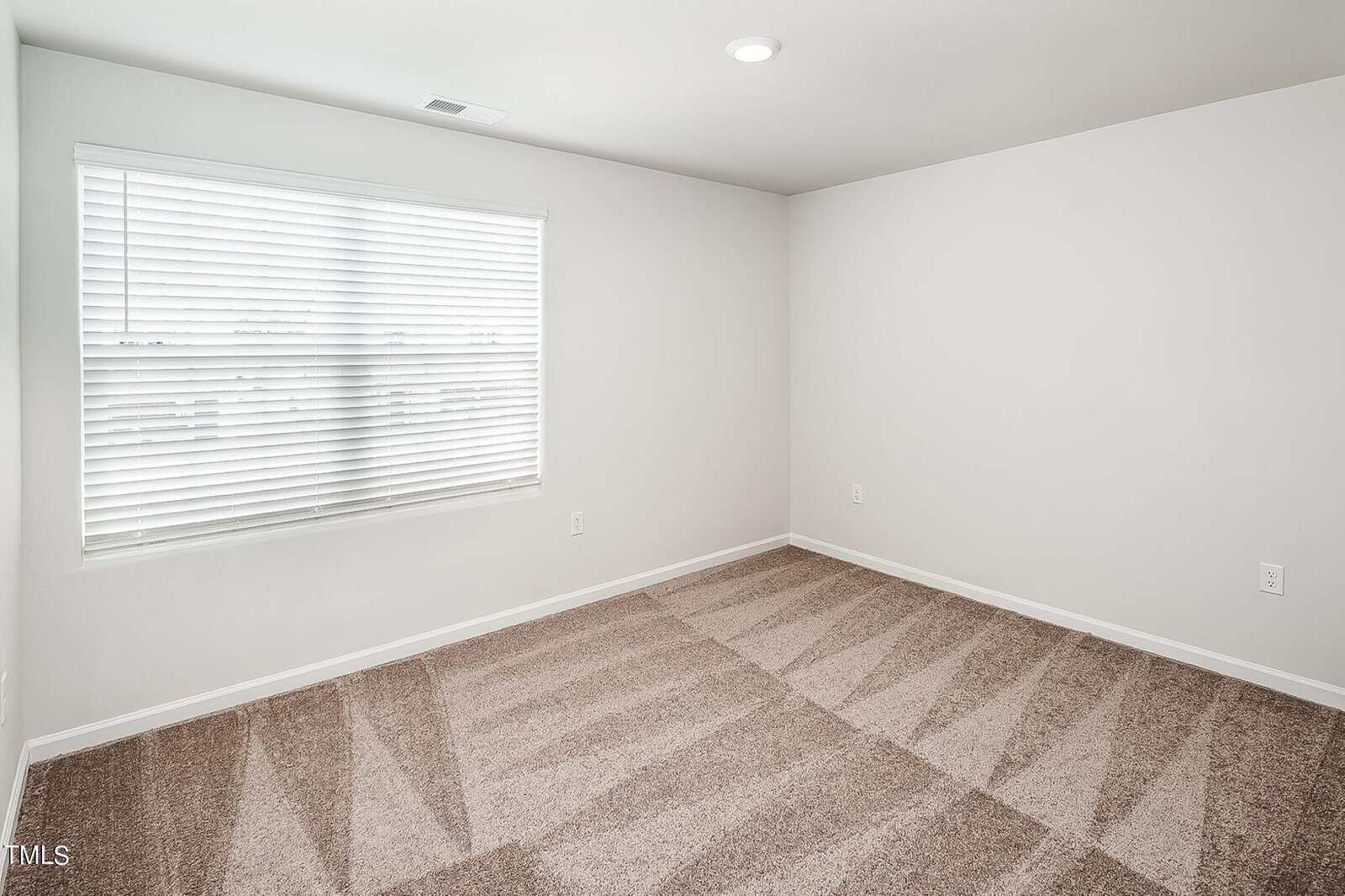 A clean, empty room with light-colored walls and a carpeted floor featuring striped vacuum lines. The room is illuminated by a ceiling light and natural light from a window with white blinds.