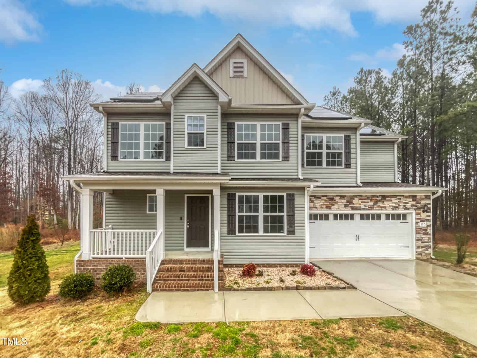 Two-story suburban house with gray siding, white trim, a covered front porch, double garage, and brick accents. The driveway and walkway are wet, with a small front garden and trees in the background under a blue sky.