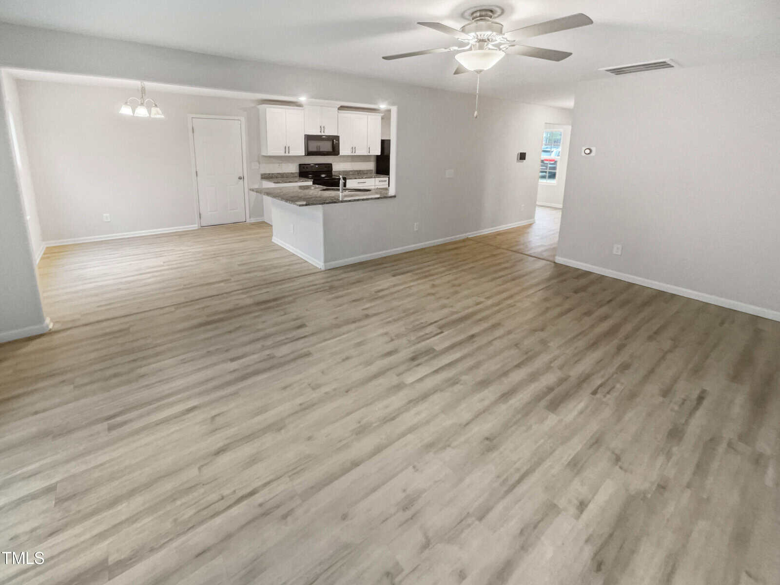Spacious open-concept living area with light wood flooring, white walls, ceiling fan, and recessed lighting. The kitchen features white cabinets, stainless steel appliances, and a bar countertop. Dining area visible to the left.