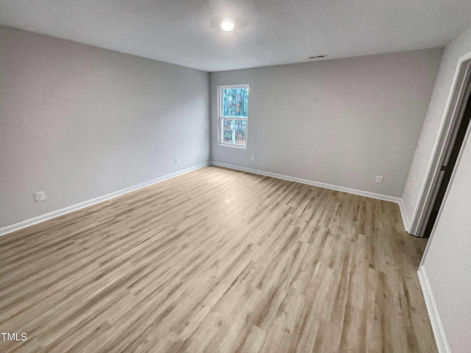 A spacious, empty room with light wood flooring, light gray walls, a single ceiling light, and a window letting in natural light. There is a doorway on the right wall.