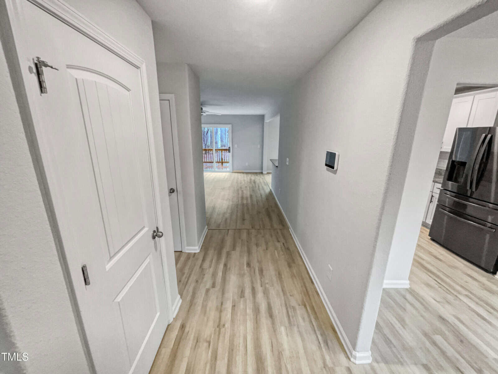 A hallway with light wood flooring leads to a bright living area with large windows. To the right, a kitchen with white cabinets and stainless steel appliances is partially visible. Walls are painted white.