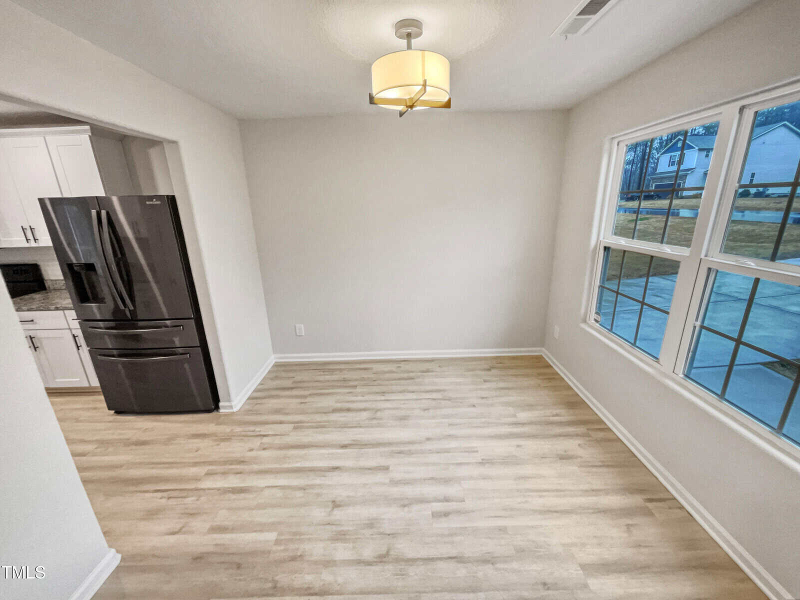 A bright, empty dining area with light wood flooring, a modern ceiling light, and large windows. A stainless steel refrigerator and part of a white kitchen are visible to the left.
