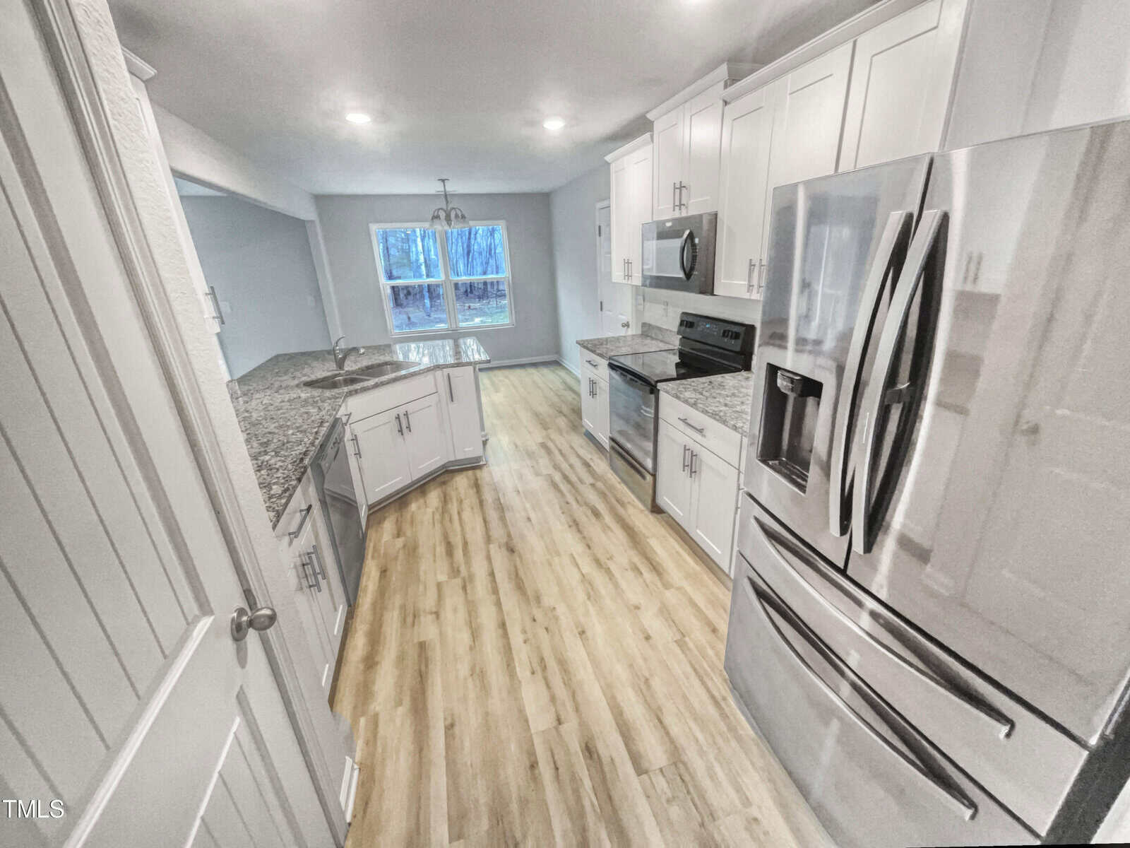 Modern kitchen with stainless steel appliances, white cabinets, granite countertops, wood-look flooring, and a large window letting in natural light at the far end of the room.