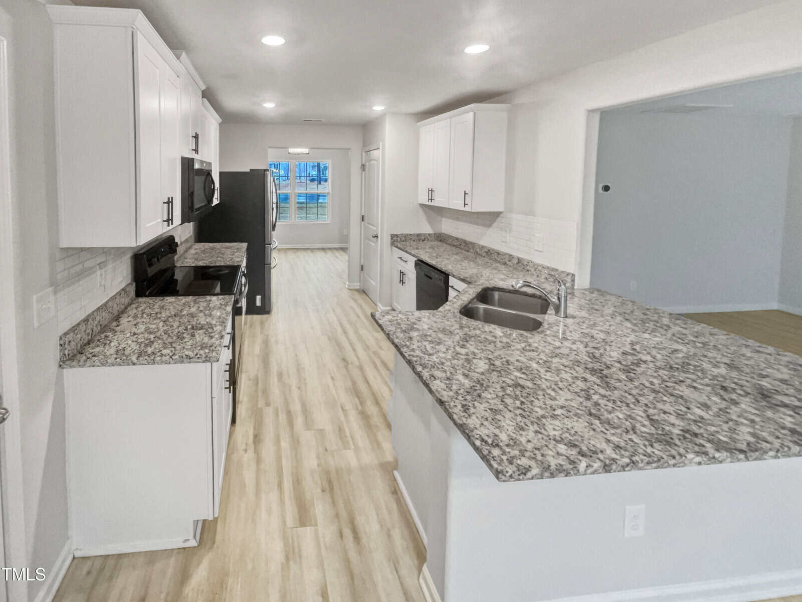 Modern kitchen with white cabinets, granite countertops, stainless steel appliances, and light wood flooring. The space is open and bright, with an open view into an adjoining room and a window in the background.
