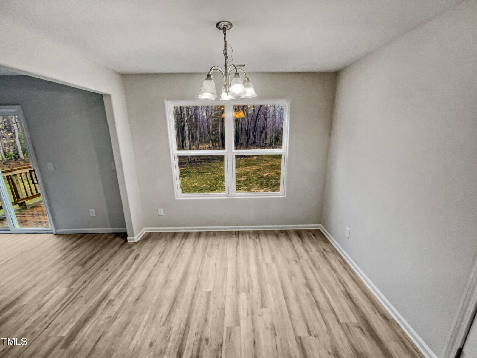 Bright, empty dining area with light wood flooring, a window showing a wooded yard, and a ceiling light fixture; an open doorway leads to a deck outside.