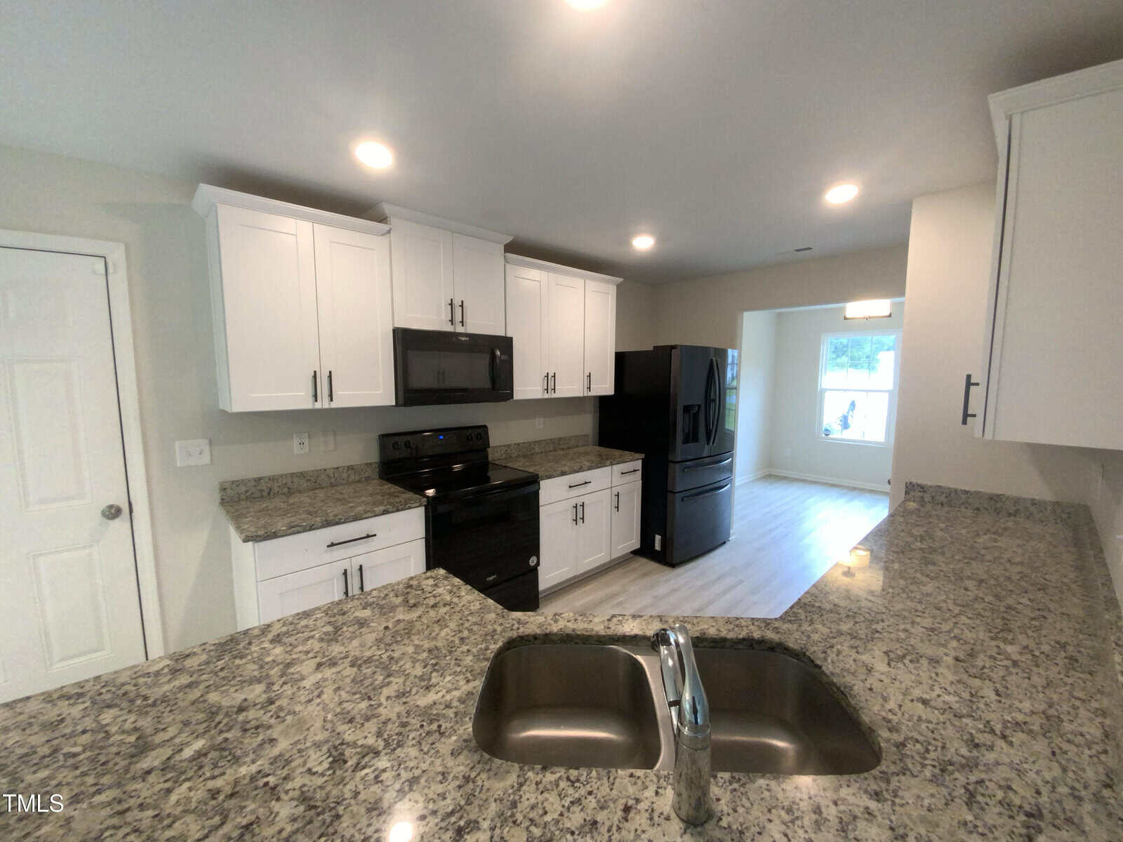 Modern kitchen with white cabinets, granite countertops, a double sink, black appliances, and recessed lighting. A window is visible in the dining area beyond the open layout.