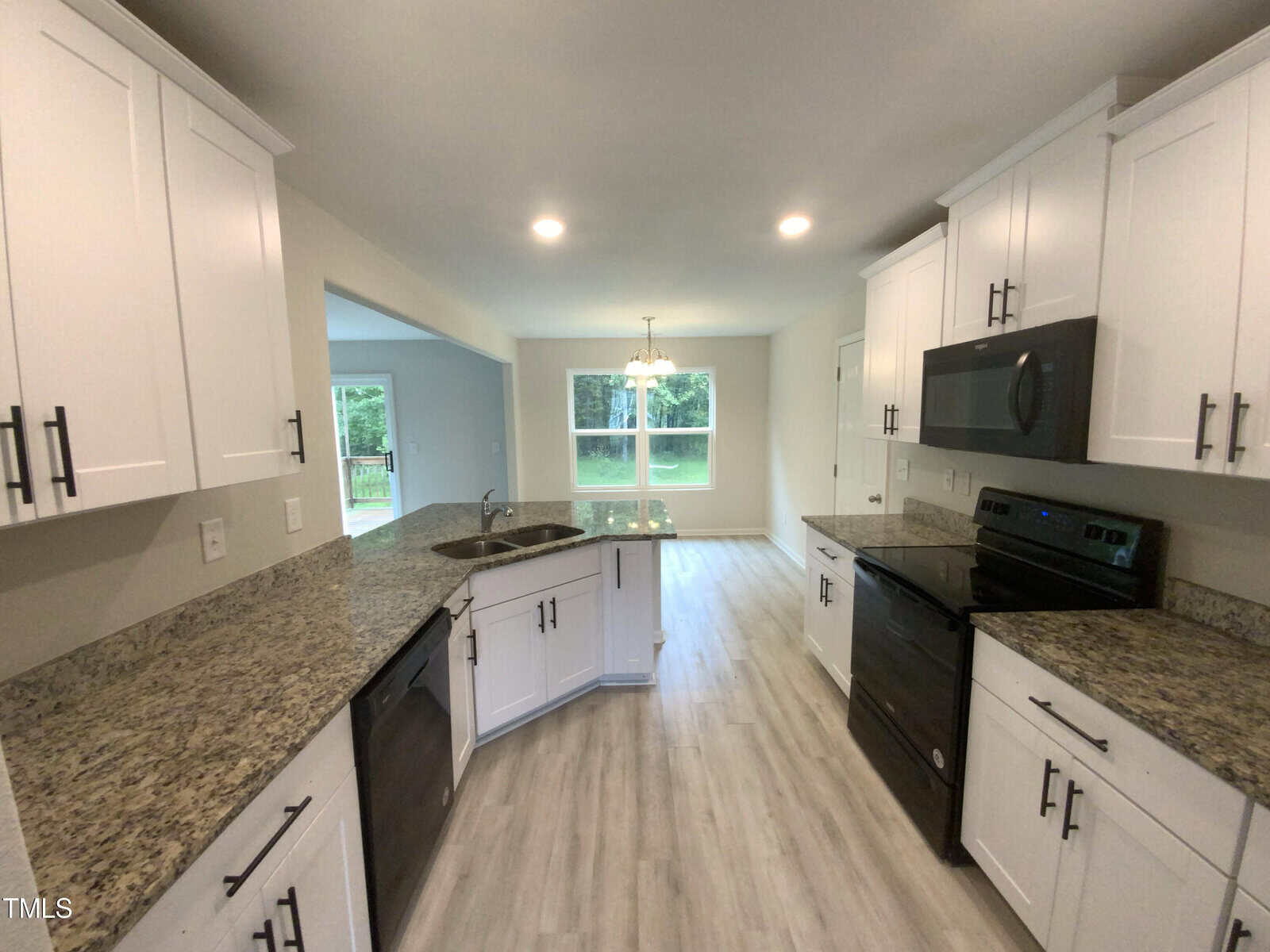 Modern kitchen with white cabinets, granite countertops, stainless steel appliances, and light wood flooring. An open layout connects to a dining area with windows overlooking greenery.