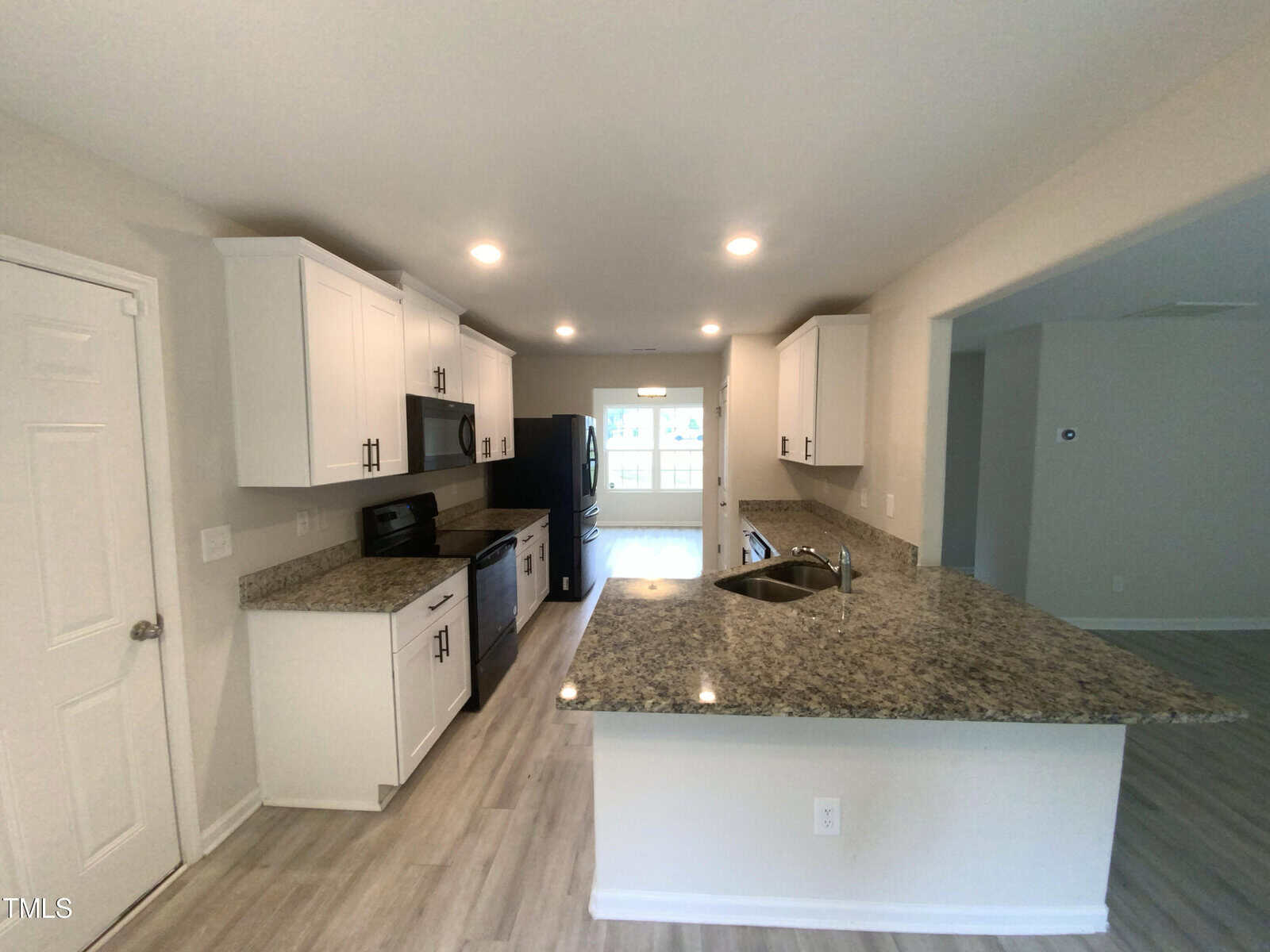 Modern kitchen with white cabinets, granite countertops, stainless steel appliances, a double sink, and wood-style flooring. Open layout leads into a bright dining area with large windows in the background.
