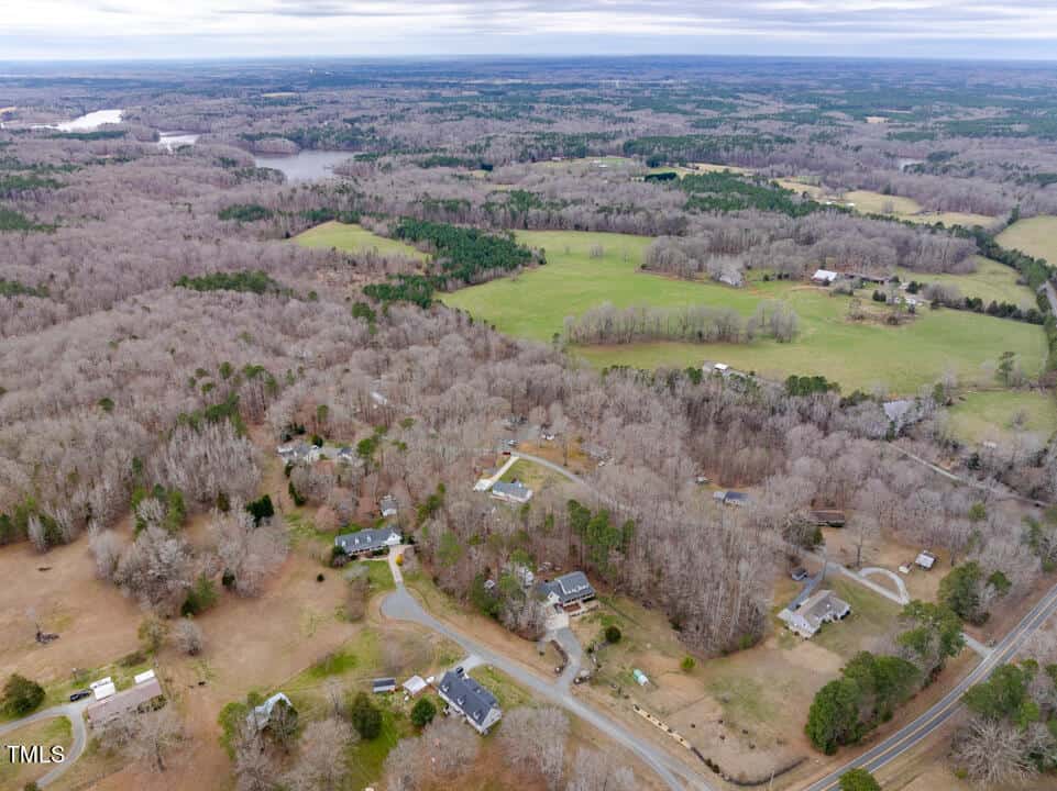 Aerial view of a rural landscape with scattered houses, winding roads, and leafless trees. Open fields and forests are visible, with a lake in the background under a cloudy sky.