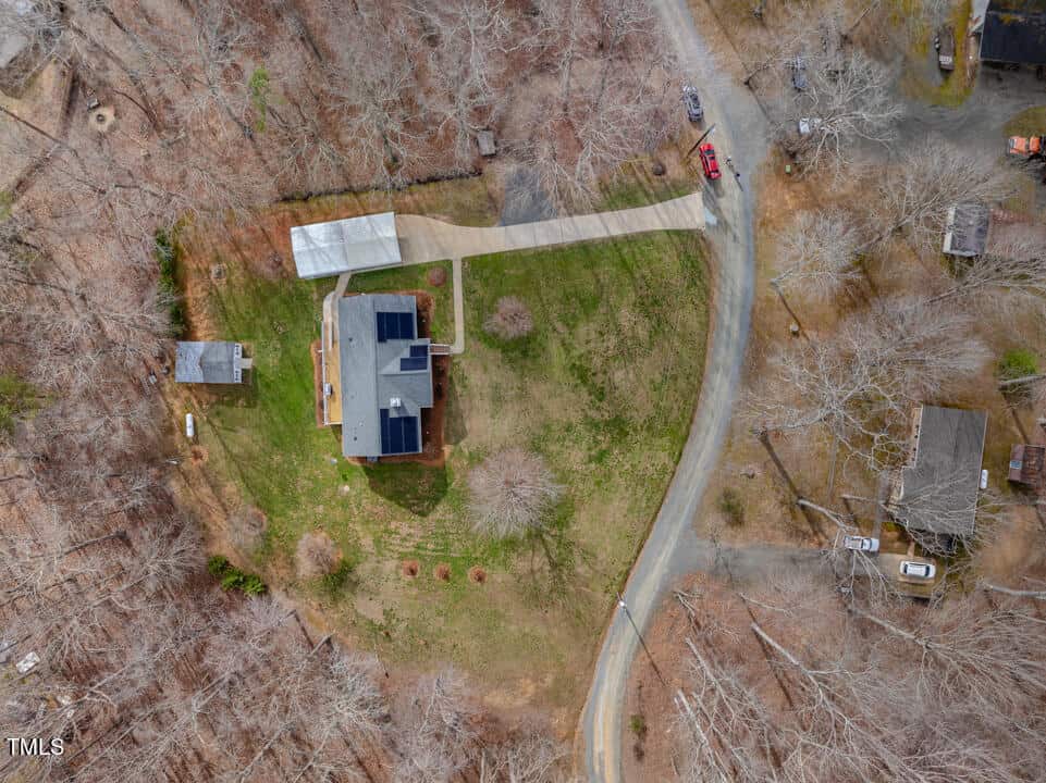 Aerial view of a rural property featuring a house with solar panels on the roof, a detached smaller building, and a long driveway. Surrounding the property is a mix of trees with barren branches, indicating a late fall or winter season.