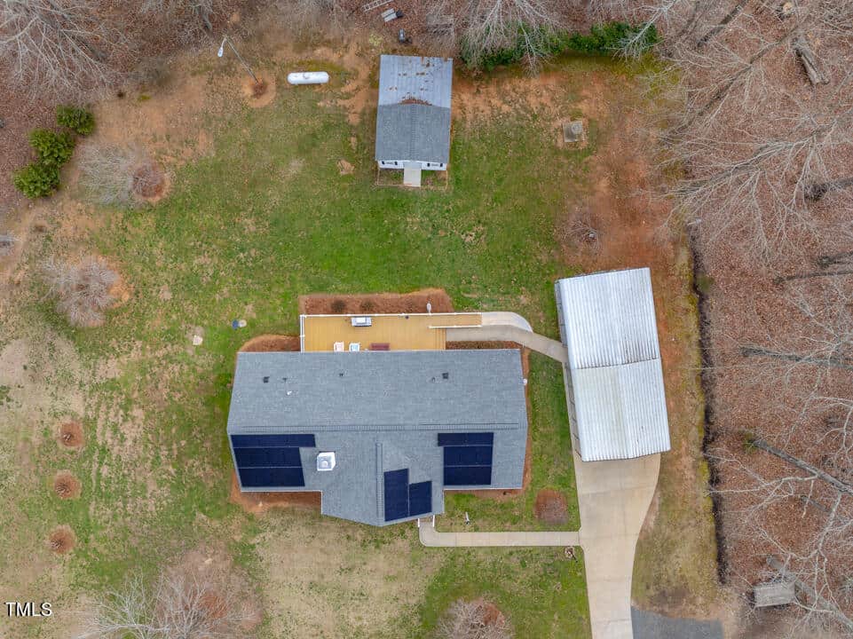 Aerial view of a property with a main house featuring a large deck and solar panels, a detached garage, and a smaller shed. The yard is spacious with scattered trees and brown grass. A driveway leads from the road to the garage.