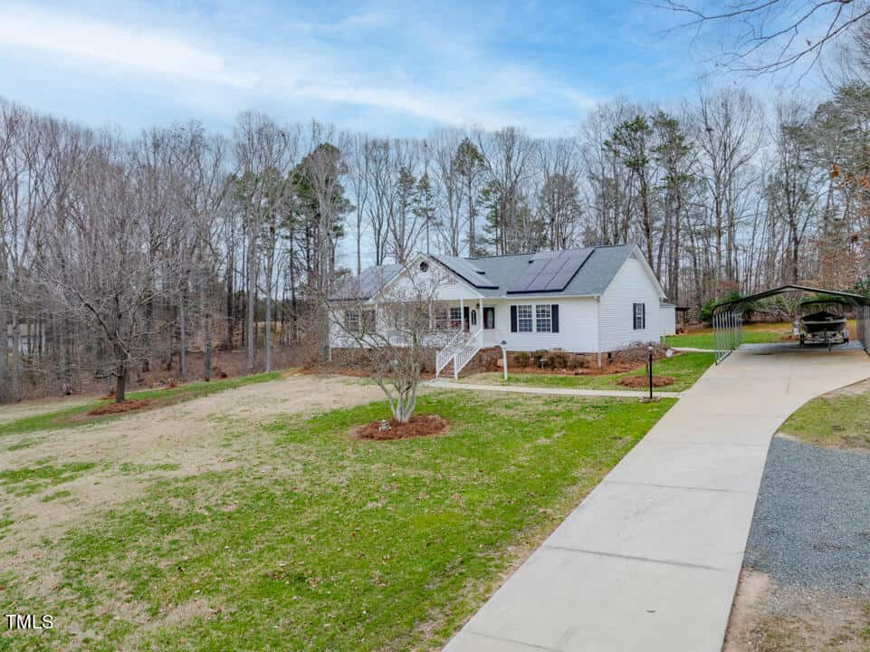 A white single-story house with solar panels on the roof sits in a clearing surrounded by trees. There is a paved driveway leading to the house and a carport. The yard has patches of grass and a few shrubs. The background features leafless trees.