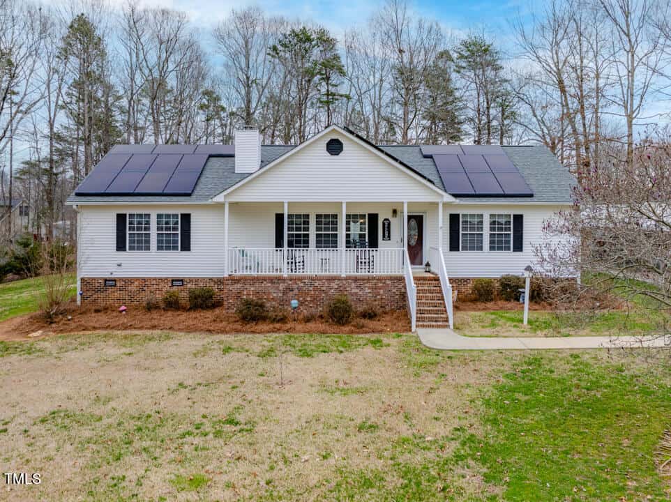 Located at 3536 Fletchers Way, this charming single-story white house features a welcoming front porch and eco-friendly solar panels on the roof. The yard, with sparse grass and bare trees, leads to the porch via a paved path, while a chimney stands proudly on the left.
