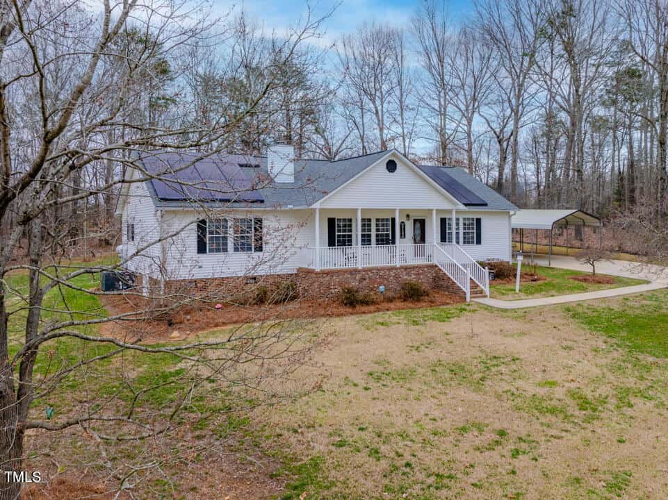 Single-story white house with a front porch, surrounded by trees and a grassy yard. Solar panels are installed on the roof. A driveway leads to a covered parking area on the side. The sky is clear, and the setting is serene.