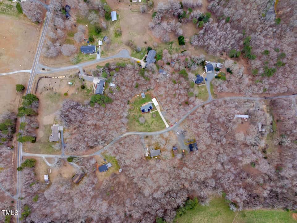 Aerial view of a rural area with several houses surrounded by trees and fields. A winding road connects the properties. The landscape is composed of a mix of open grassy areas and dense woods, with hints of winter foliage.