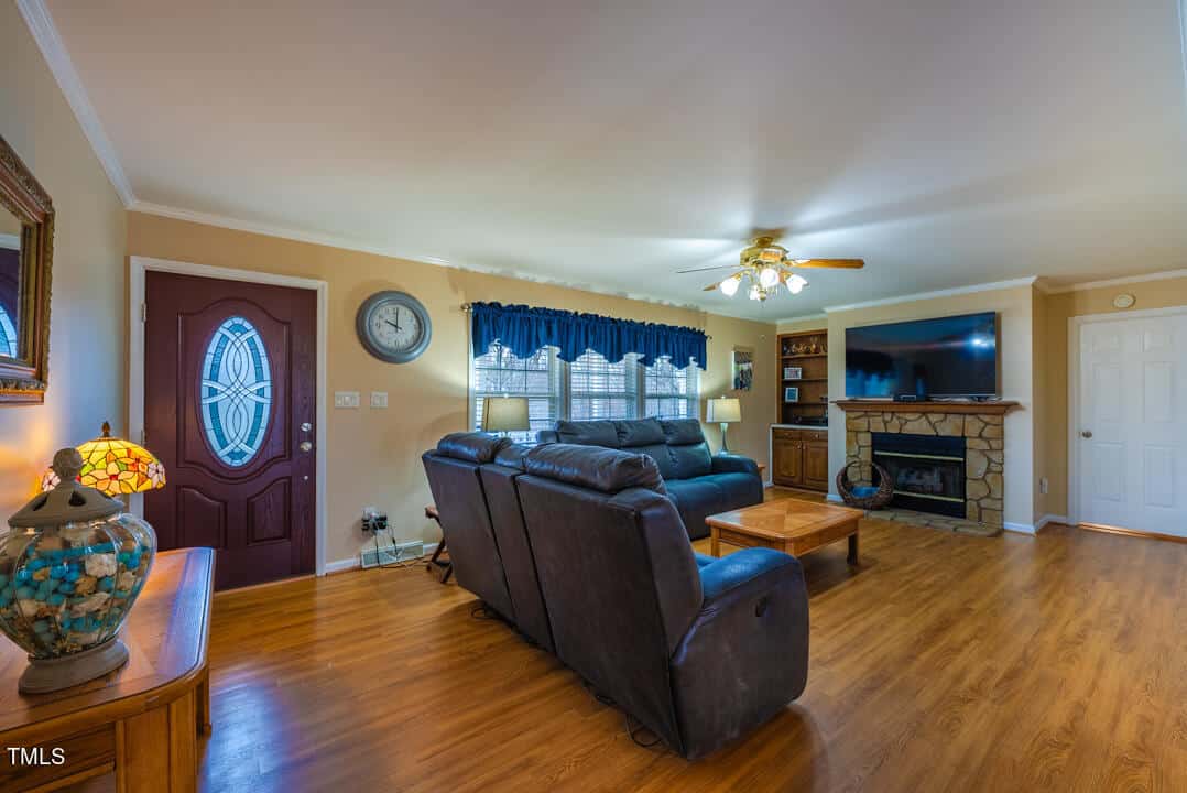 Cozy living room with wooden floor, dark leather sofas, and a stone fireplace. A large TV is mounted on the wall. A round clock hangs above a window with blue curtains. A decorative lamp and a round mirror adorn a table near the entrance.
