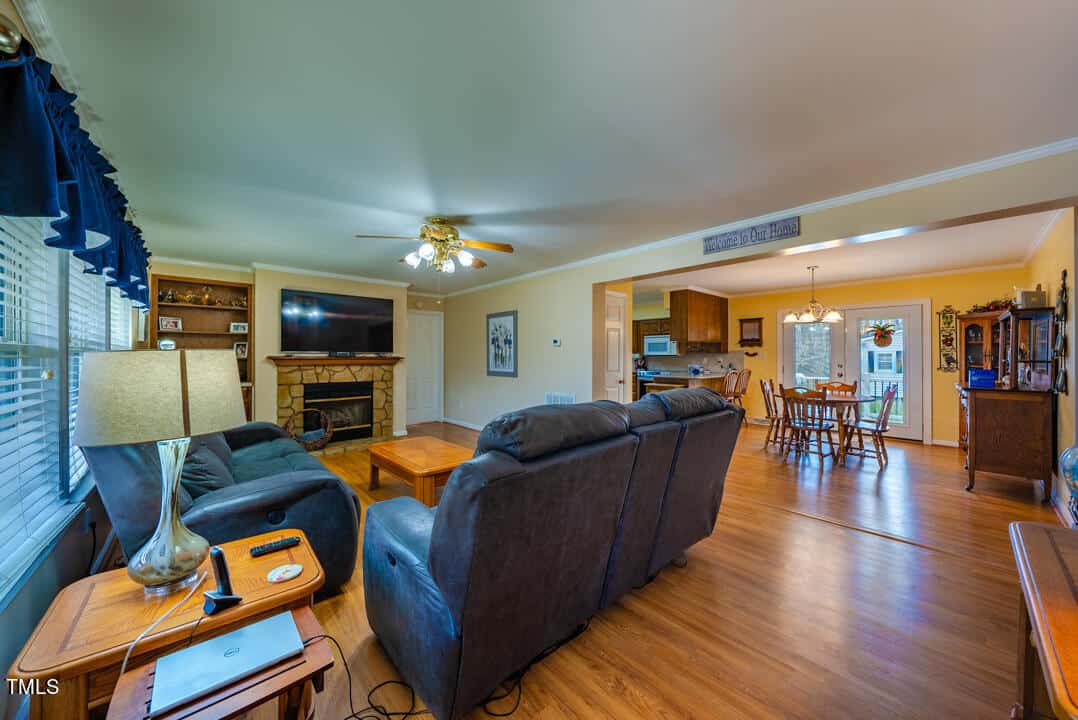 Spacious living room with a stone fireplace, large TV, and two dark sofas. The area is brightly lit with a ceiling fan. In the background, a dining area is visible, leading to a kitchen with wooden cabinets. Hardwood floors throughout.