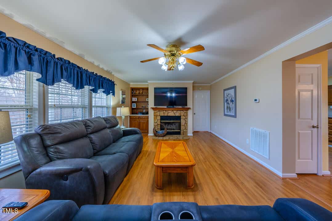 A spacious living room with a ceiling fan, large windows with blue curtains, a stone fireplace with a TV above, and comfortable dark blue seating. The room features wooden floors and a wooden coffee table at the center.