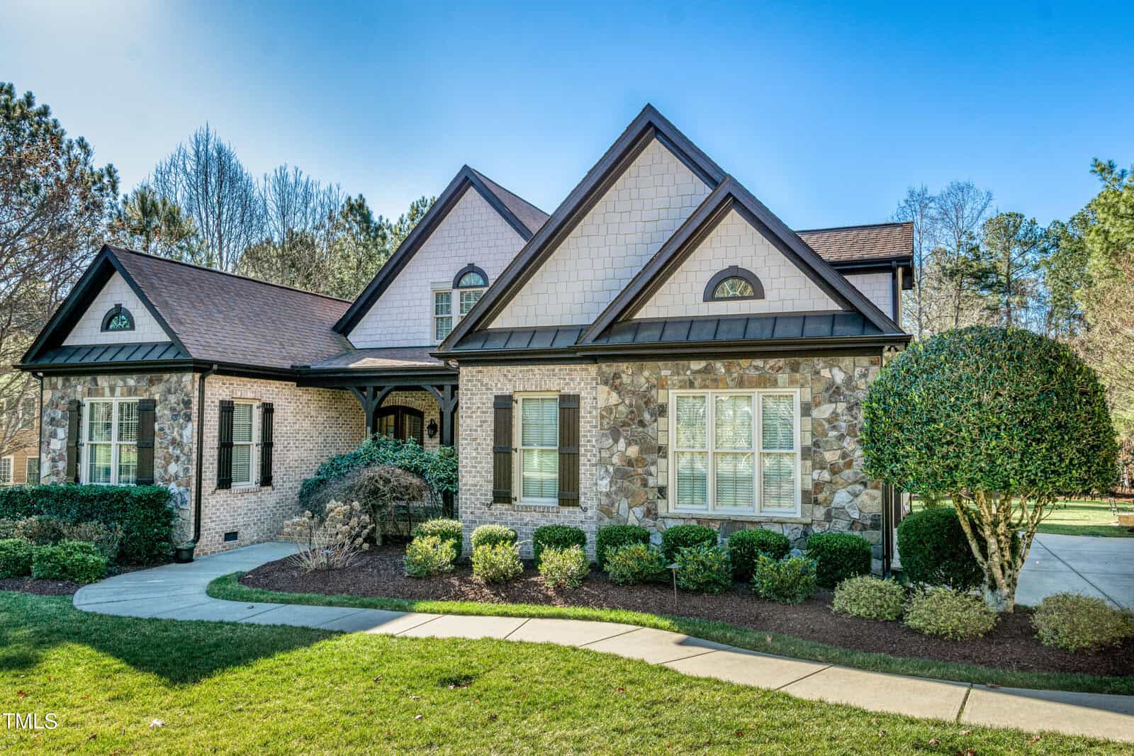 A charming house with a stone and brick exterior at 3009 Krogen Court, featuring a gabled roof and large windows. The front yard in Creedmoor is landscaped with neatly trimmed bushes and a curved walkway leading to the entrance. Trees and blue sky are visible in the background.
