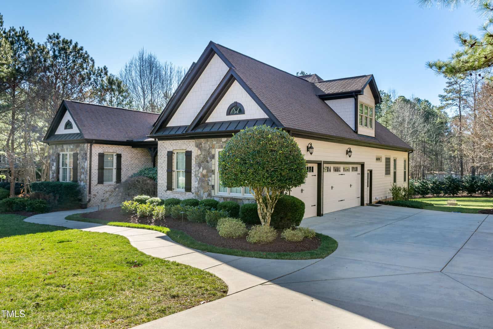 A suburban house with a brick and stone facade features a neatly manicured lawn and driveway. The home has a peaked roof, light siding, and two garage doors. Tall trees surround the property, providing a natural backdrop.