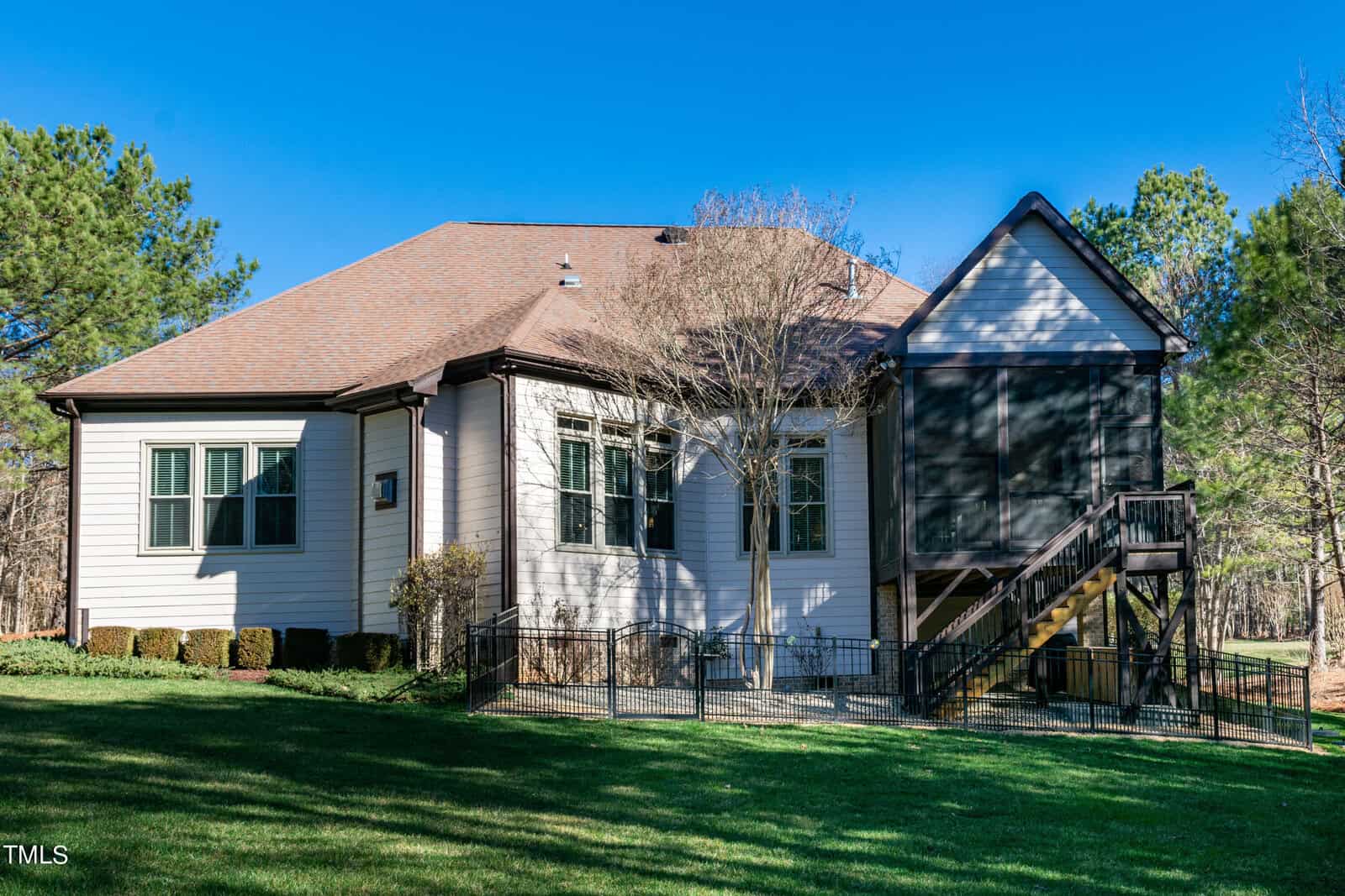 A single-story white house with a brown roof, surrounded by trees. It features a screened porch, fenced yard, and wooden staircase leading to a side entrance. The yard is well-maintained with green grass and clear blue sky overhead.