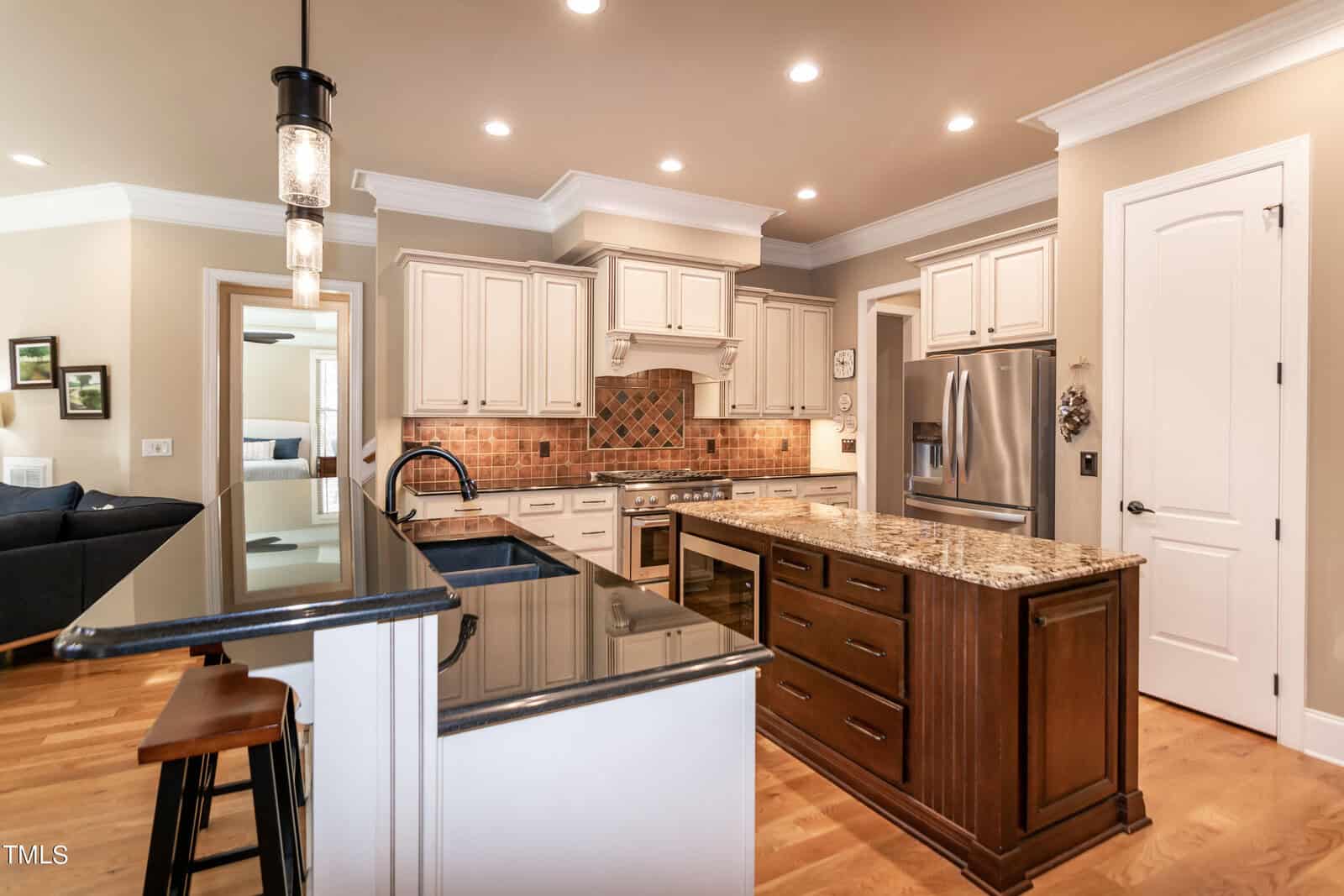 Spacious kitchen with white cabinets, a central island with a granite countertop, and a double sink. Stainless steel appliances, including a refrigerator and oven, are present. Pendant lights hang above the island, and wood flooring completes the look.
