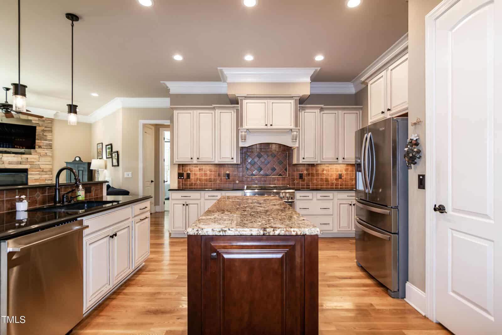 A spacious kitchen with hardwood floors features a large island with a granite countertop. Surrounding are stainless steel appliances, white cabinets, and a tile backsplash. Pendant lights hang over the island, and a door is visible on the right.