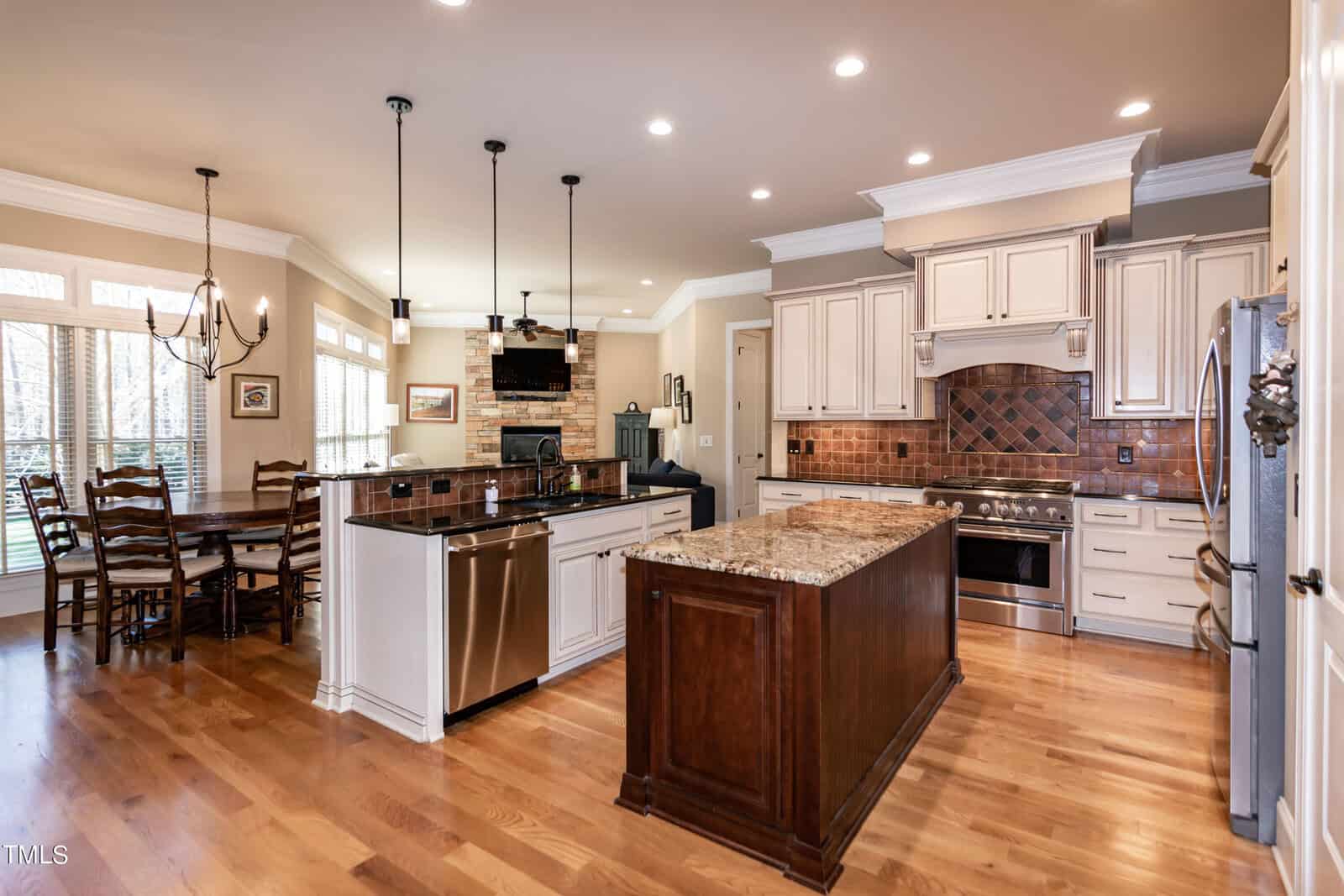 A spacious kitchen with wooden floors features a central island with granite countertops, stainless steel appliances, and cream-colored cabinets. A dining area with a table and chairs is seen on the left, illuminated by natural light from large windows.