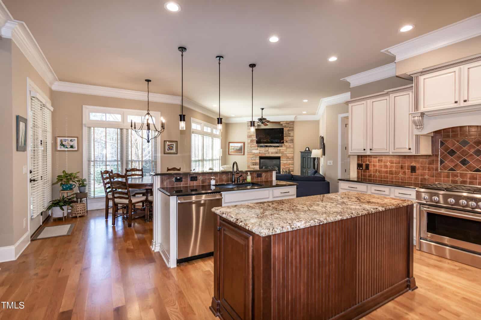A spacious kitchen with wood flooring, granite countertops, and white cabinets. It features an island, stainless steel appliances, and a sink. Pendant lights hang above the island, and a dining area is visible in the background near large windows.