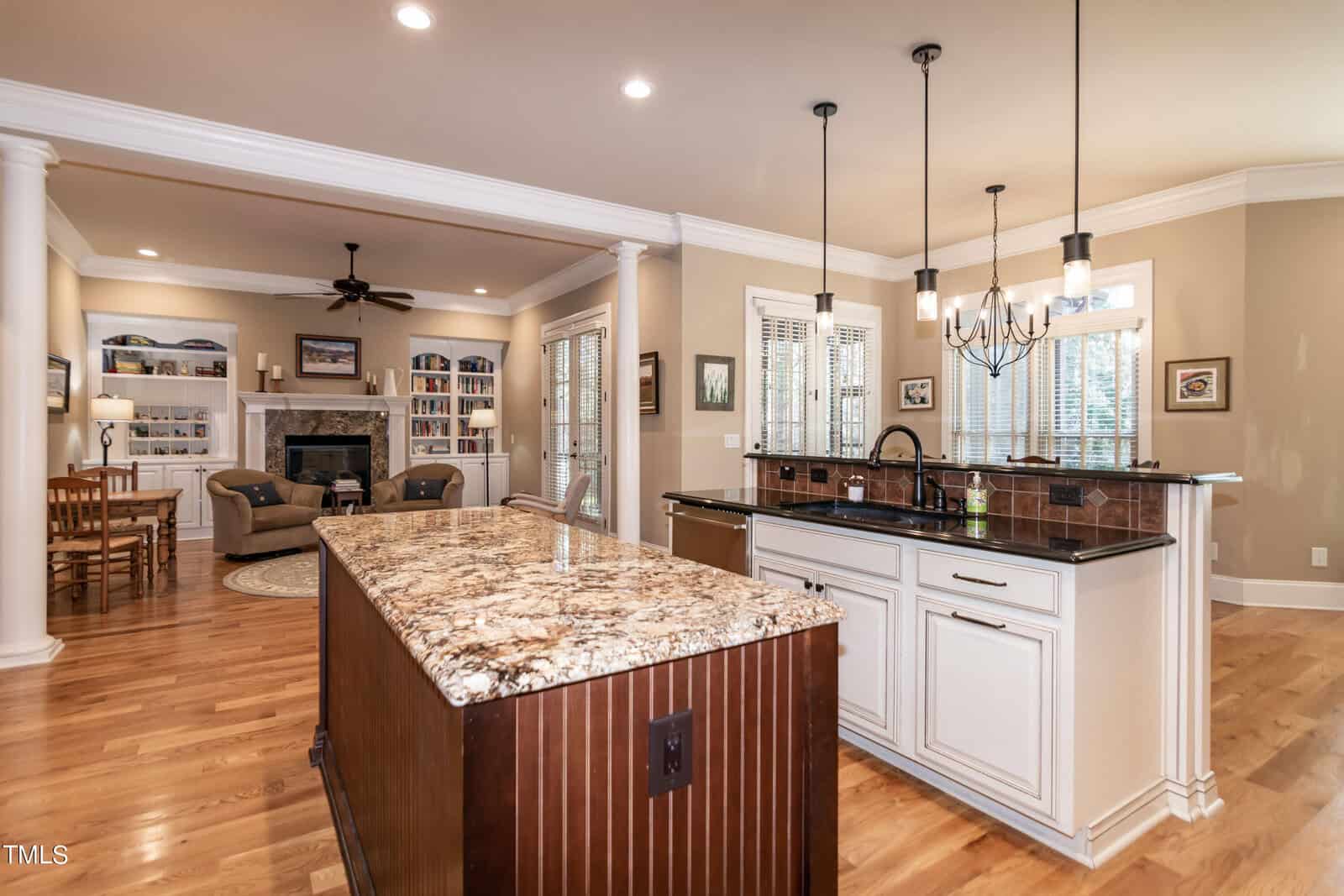 Spacious kitchen with granite island, white cabinets, and dark fixtures. Open layout flows into living area with a fireplace, built-in shelves, and wood floors. Dining table visible in the corner. Pendant lights hang above the island.