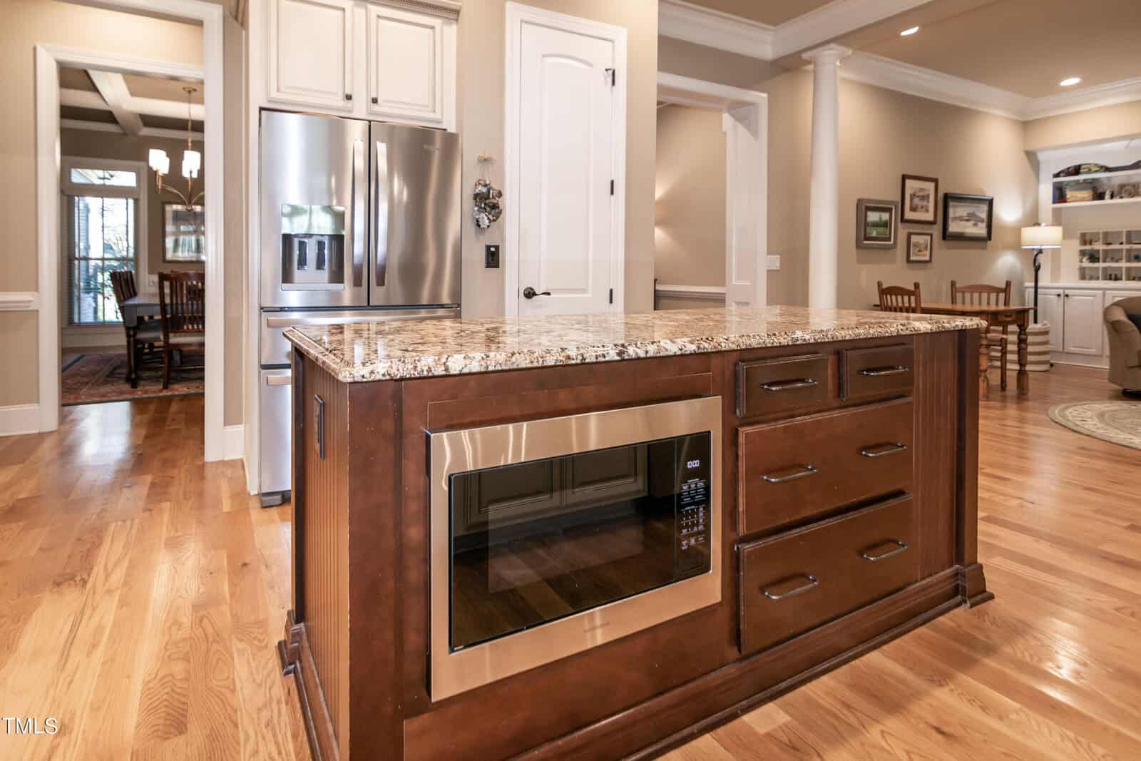 A kitchen with wooden floors and a central island featuring a microwave and drawers. Stainless steel refrigerator and a dining area are visible in the background, along with framed pictures on the walls.