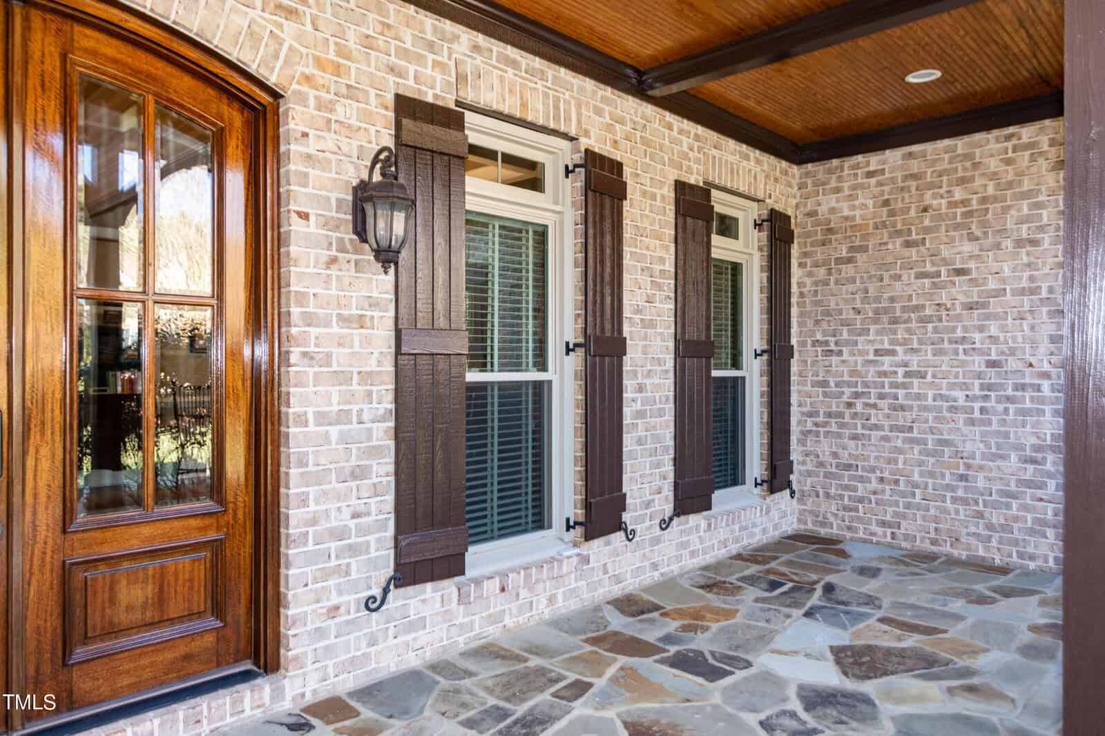 Brick house exterior with three windows featuring dark wooden shutters, a polished wooden door with glass panels, and a stone-tiled porch. A classic outdoor lantern is mounted on the wall beside the door under a wooden ceiling.