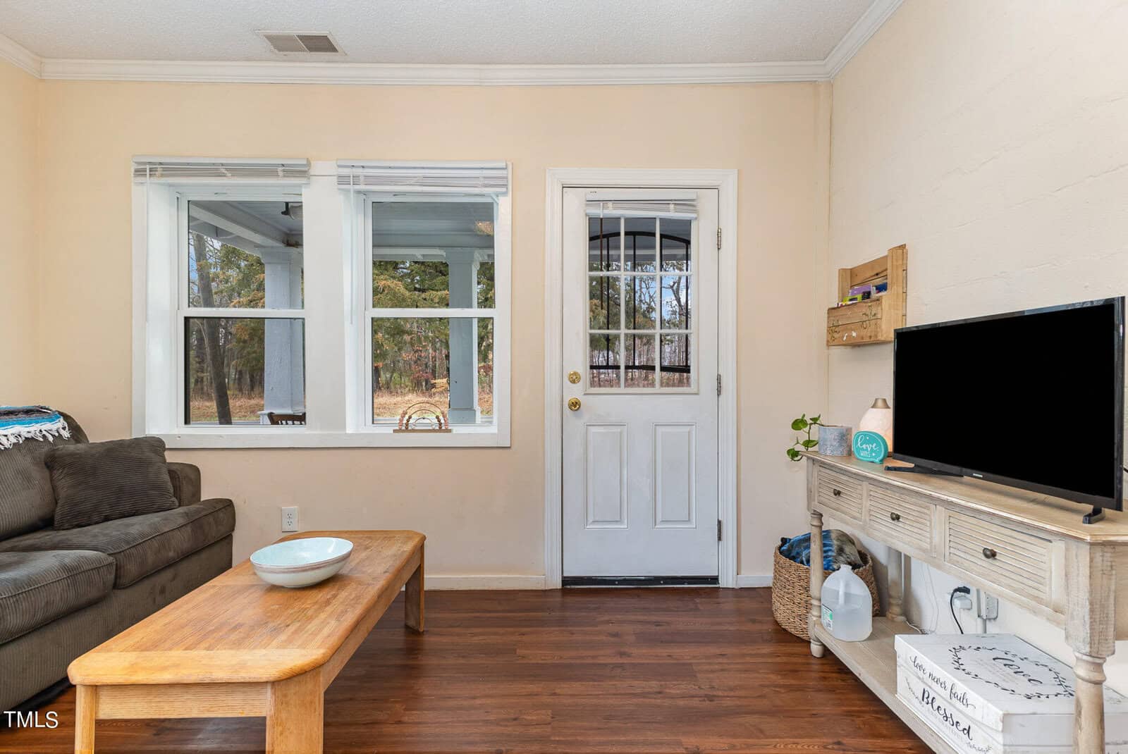 A cozy living room featuring a dark wooden floor, tan walls, two windows with white trim, and a glass-paneled door. furnishings include a sofa, a wooden coffee table, and a tv on a small console.