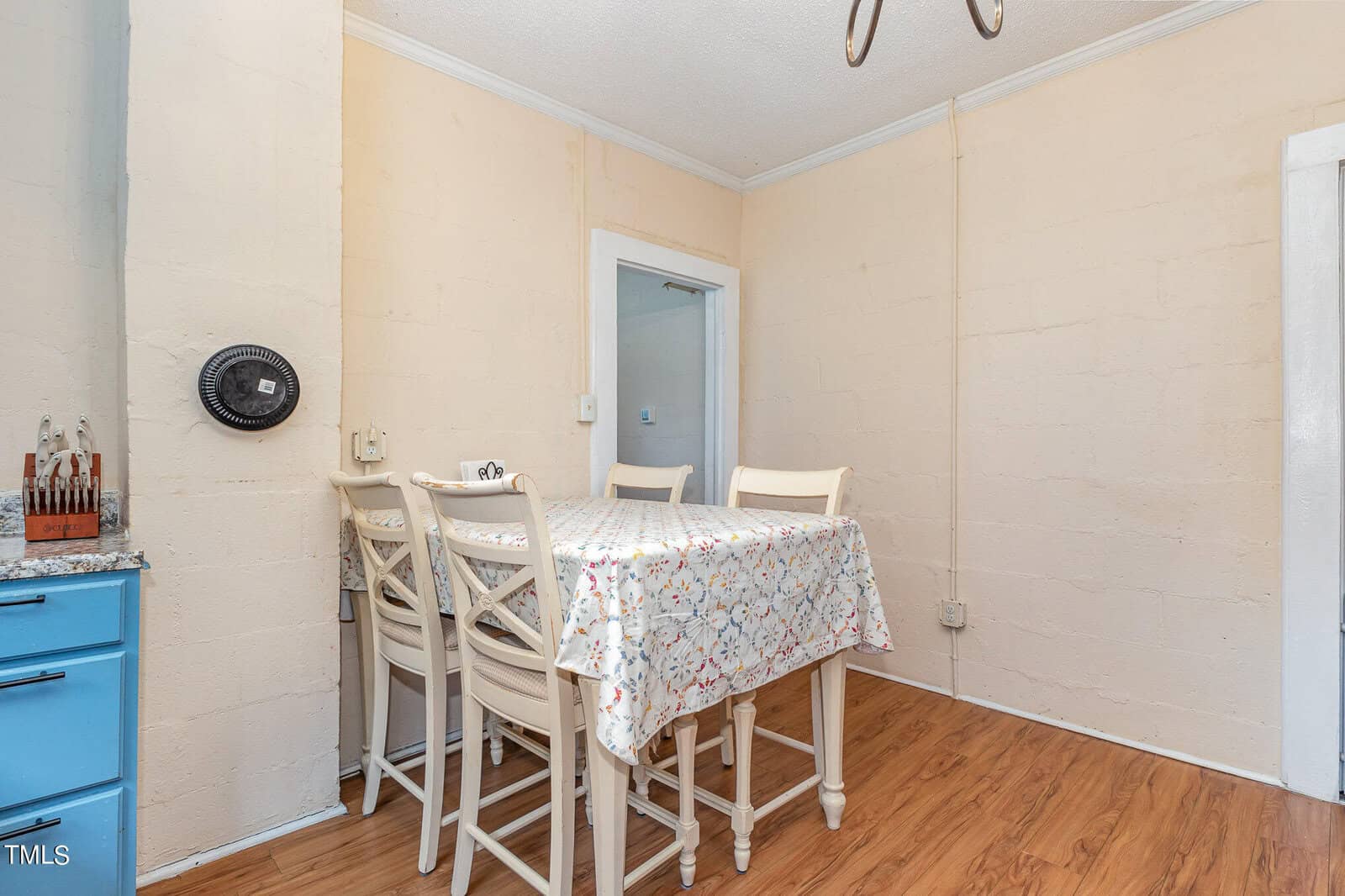 A small dining area with a wooden table covered by a floral tablecloth and four white chairs. the room features pale yellow walls, wood flooring, a wall-mounted fan, and a view into another room through a doorway.