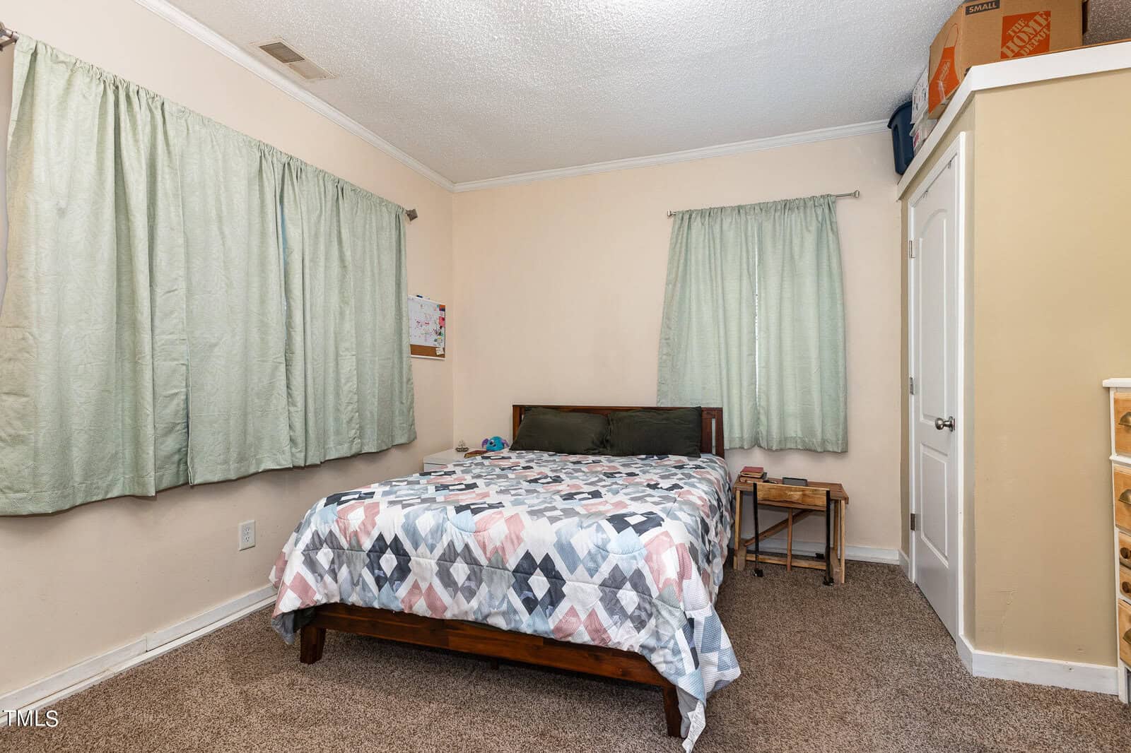 A modestly furnished bedroom with a twin bed covered in a geometric-patterned comforter, pale green curtains, a small wooden bedside table, and a closed door on the right. shelves with items are visible in a corner.