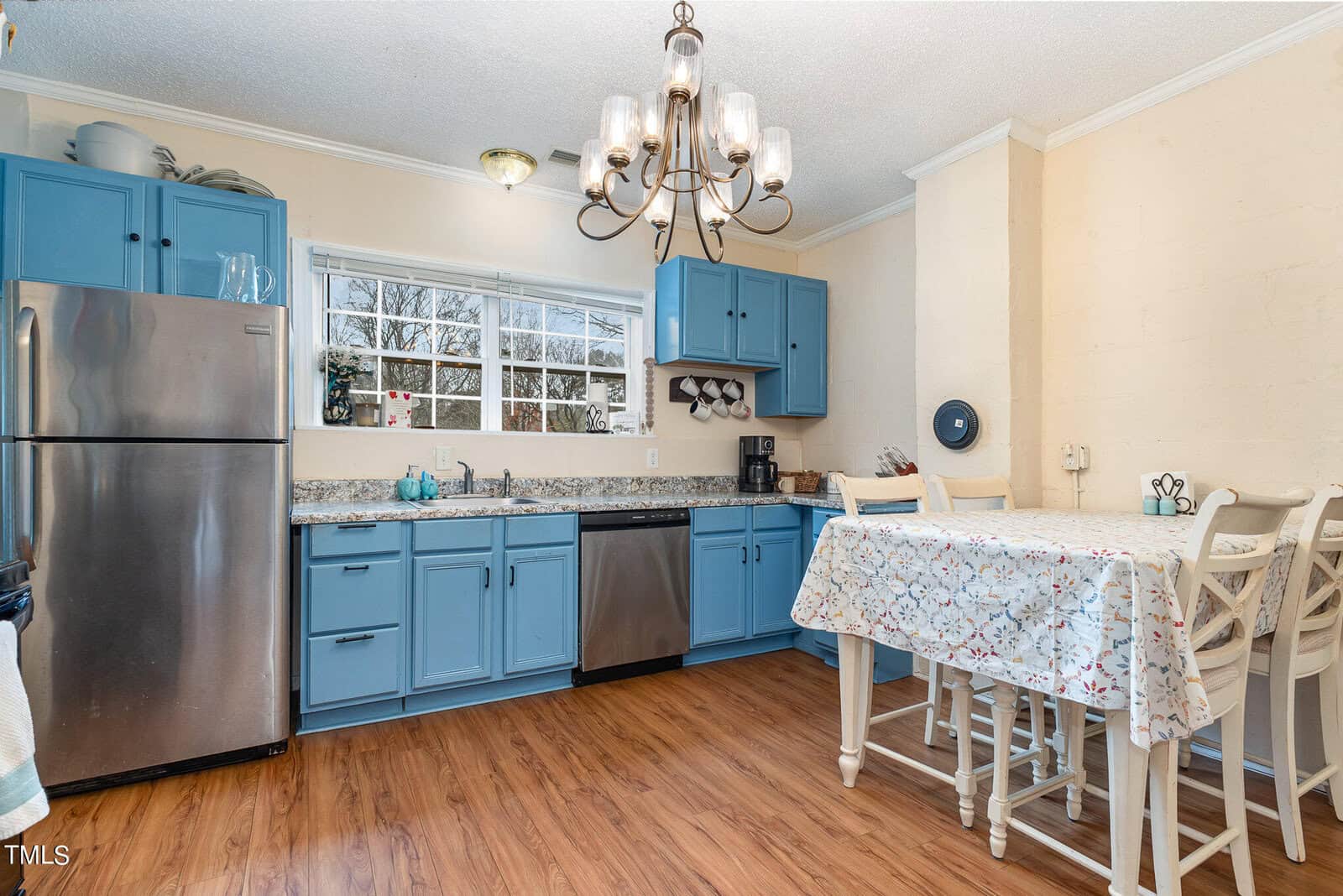A kitchen with blue cabinets, stainless steel appliances, and a wooden dining table set under a chandelier, with a window overlooking the yard.