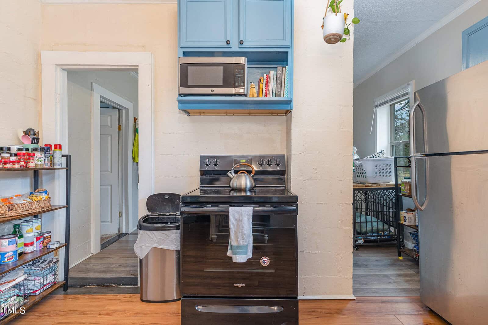 A small, cluttered kitchen showing a black stove, microwave above, and blue cabinets filled with books. to the left, an open doorway leads to another room; a white refrigerator and shelves with items are visible.