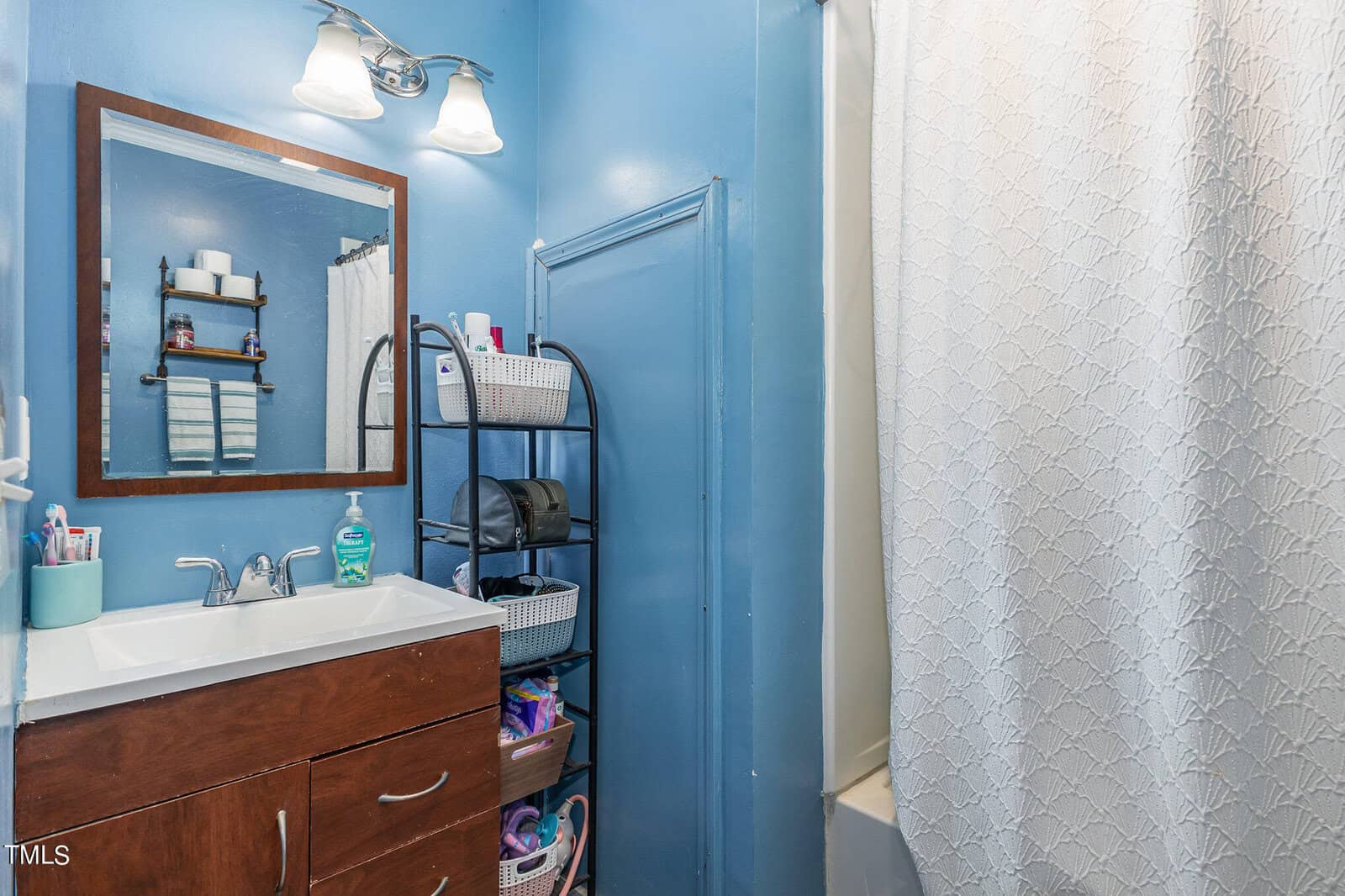 A small bathroom featuring a wooden vanity with a white sink, a framed mirror above it, a metal storage rack with toiletries, and blue walls. a white patterned shower curtain is partially visible on the right.