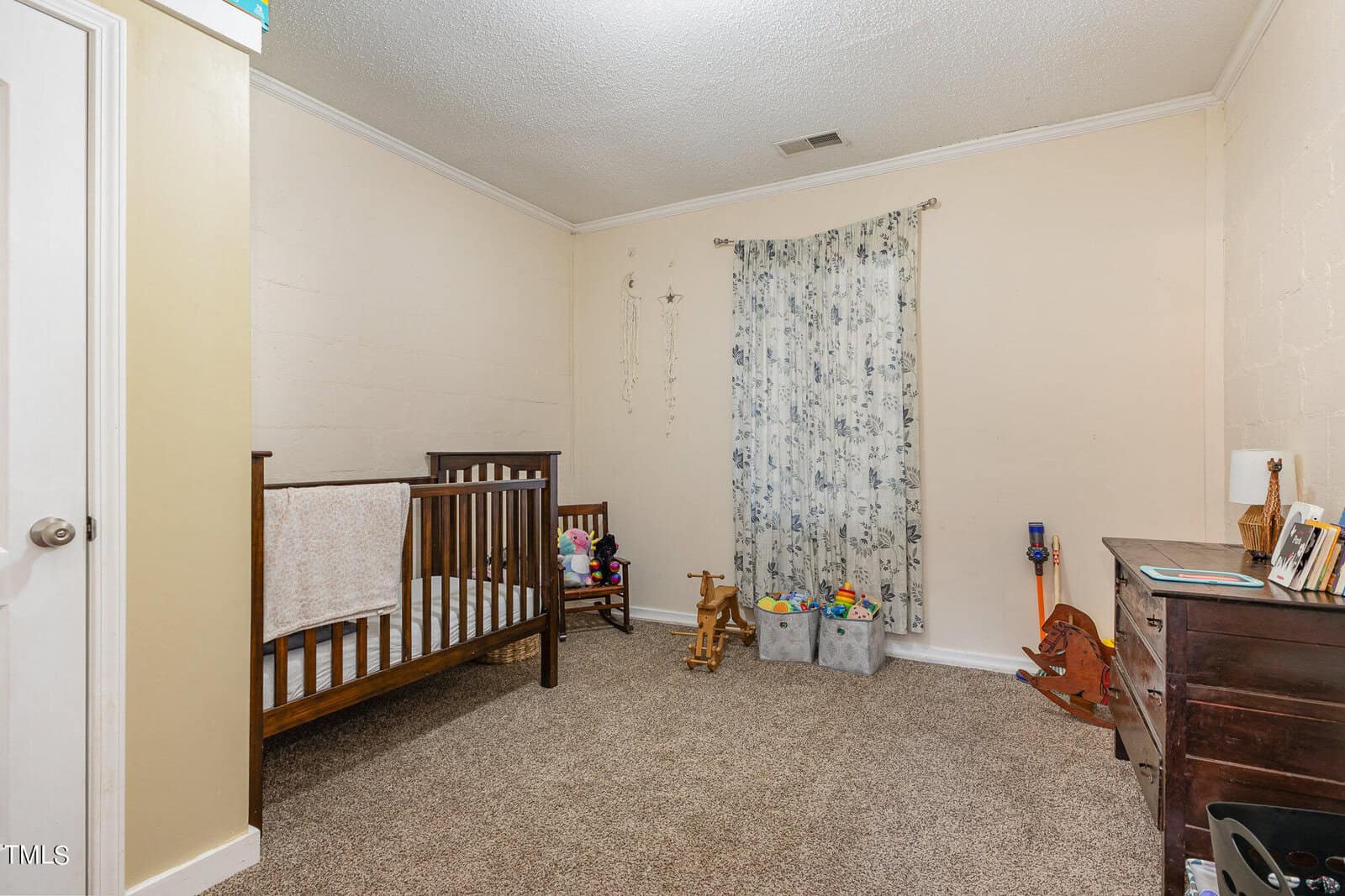 A modestly furnished nursery with a wooden crib, colorful toys, a small desk, and a window covered with floral curtains. the room features carpet flooring and white walls.