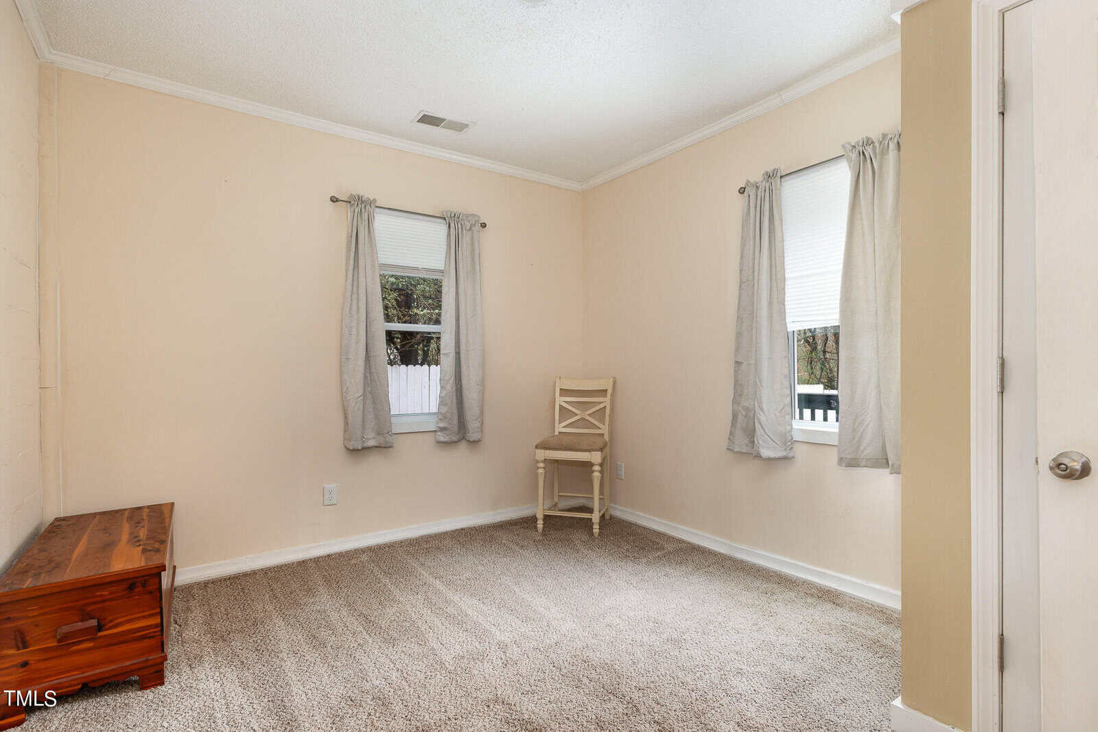 A modestly sized room featuring beige walls and a shaggy beige carpet. the room includes a single wooden chair by two curtained windows and a wooden chest near the entrance. the atmosphere is plain and unadorned.