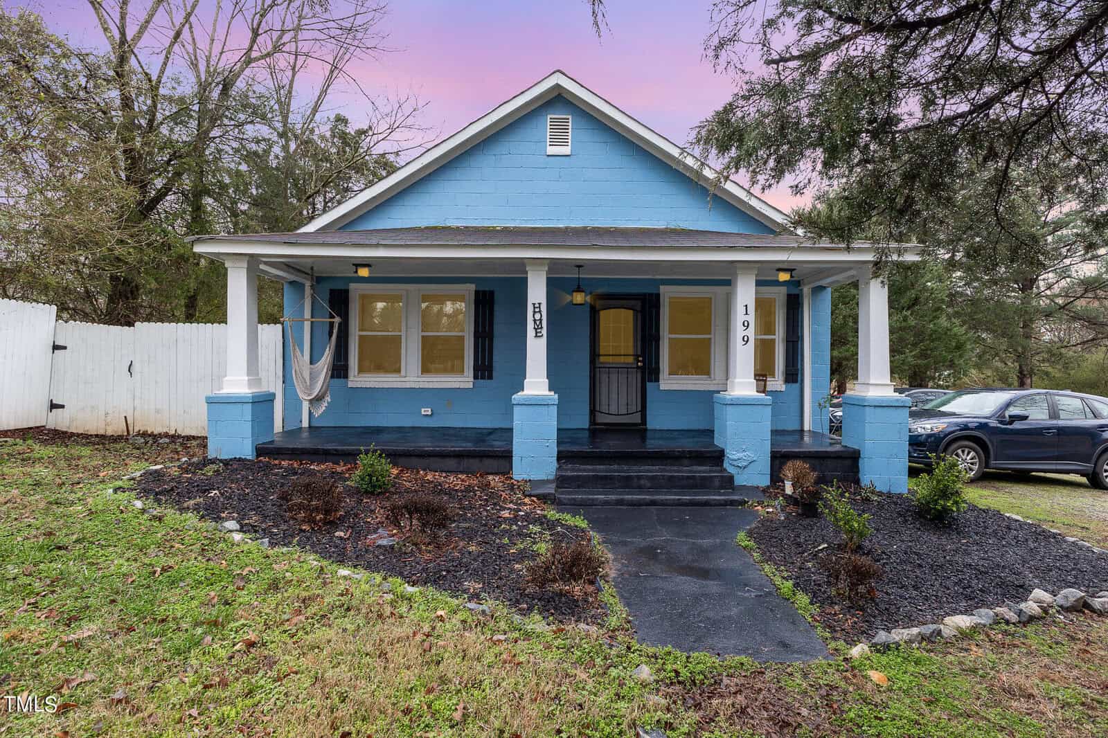 A blue single-story house with a covered front porch, black shutters, and a newly landscaped front yard. a black car is parked to the side on a driveway.