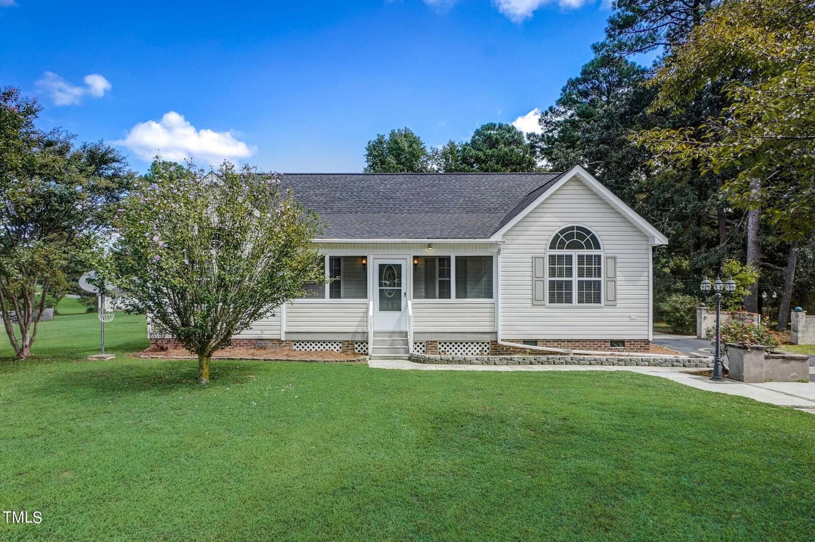 Nestled in the charming area of Franklinton, a small, single-story white house with a dark gray roof sits at 2512 Wes Sandling Road. Surrounded by verdant grass and trees, a concrete pathway guides visitors to its inviting screened front porch under a sky dotted with clouds.