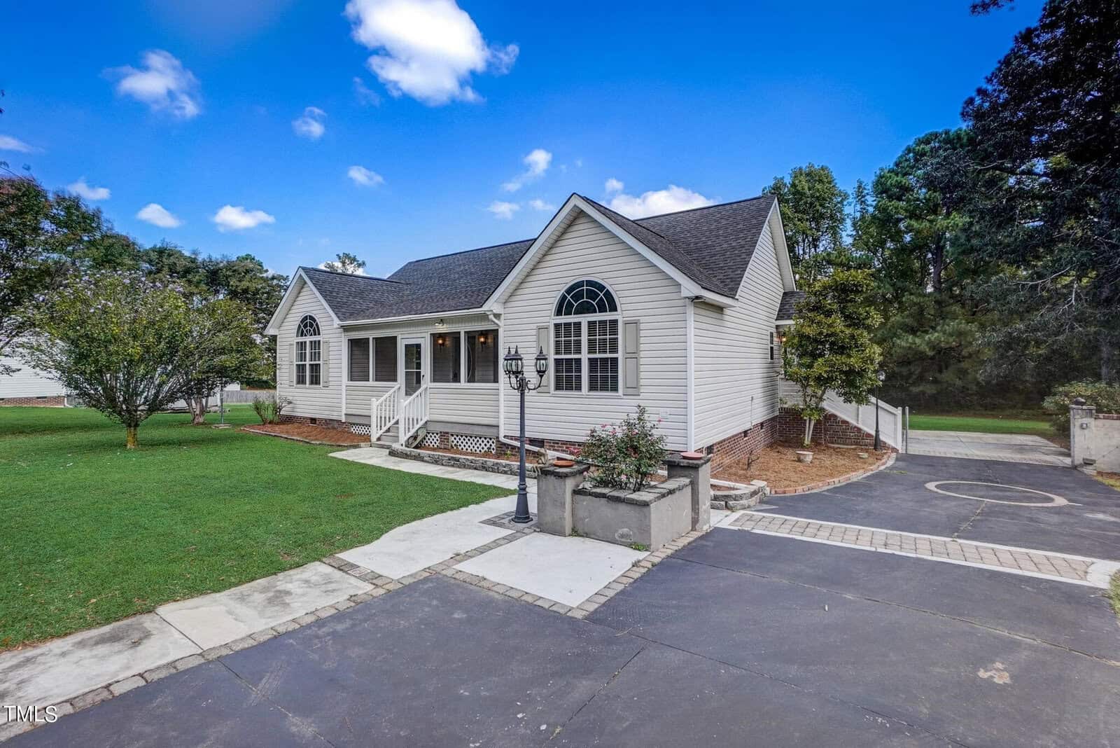 A single-story house with white siding and a dark shingled roof. It features arched windows and a small front porch. The front yard has green grass and a paved driveway. The sky is clear with a few clouds.