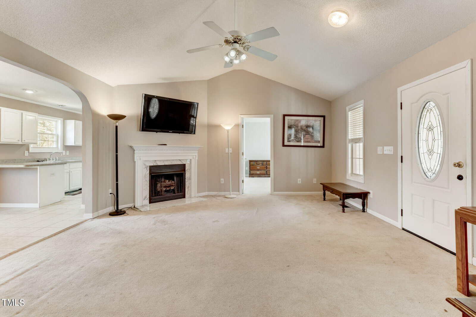A spacious living room with beige carpet, a white fireplace, and a wall-mounted TV. It features an arched doorway leading to a bright kitchen and has a ceiling fan. A decorative front door with windows is on the right, and light pours in from a window.