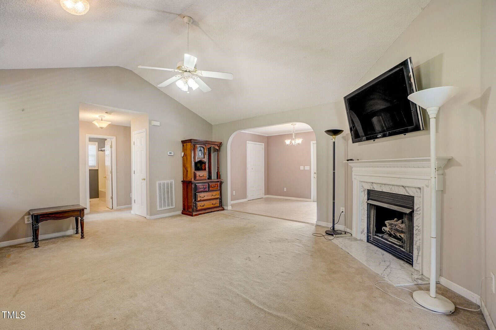 Spacious living room with beige carpet and neutral walls. Features a wall-mounted TV, white marble fireplace, ceiling fan, archway leading to another room, and wooden cabinet. Lamp stands and a small wooden table accent the space.