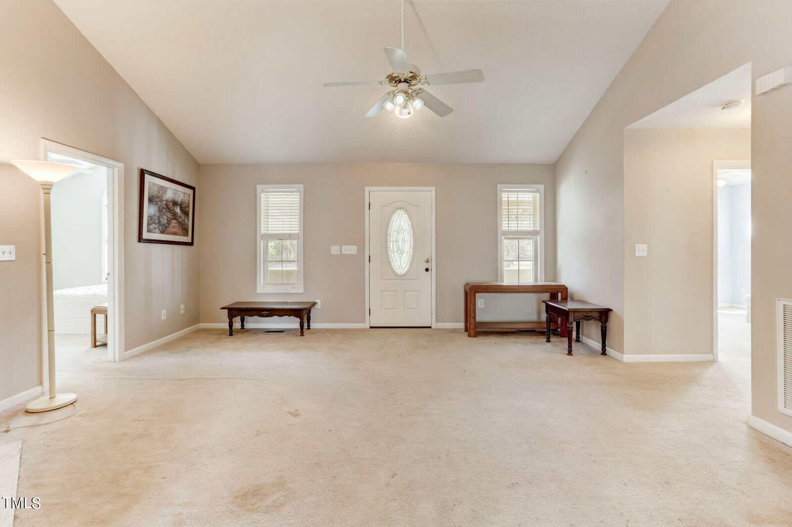 A beige living room with a high ceiling features an oval glass door at the center. Two small benches are against the walls, and a ceiling fan is above. A floor lamp stands in the left corner, and framed artwork is on one wall. Natural light illuminates the space.