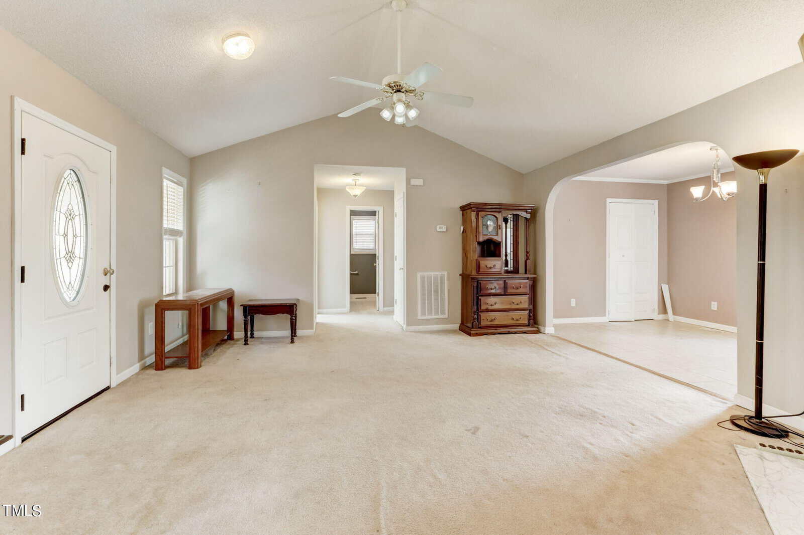 A spacious living room with beige carpet and gray walls, featuring a ceiling fan with lights. On the left, there's a round window on the door and a small wooden table. The room opens into a dining area with a chandelier and a white interior door.