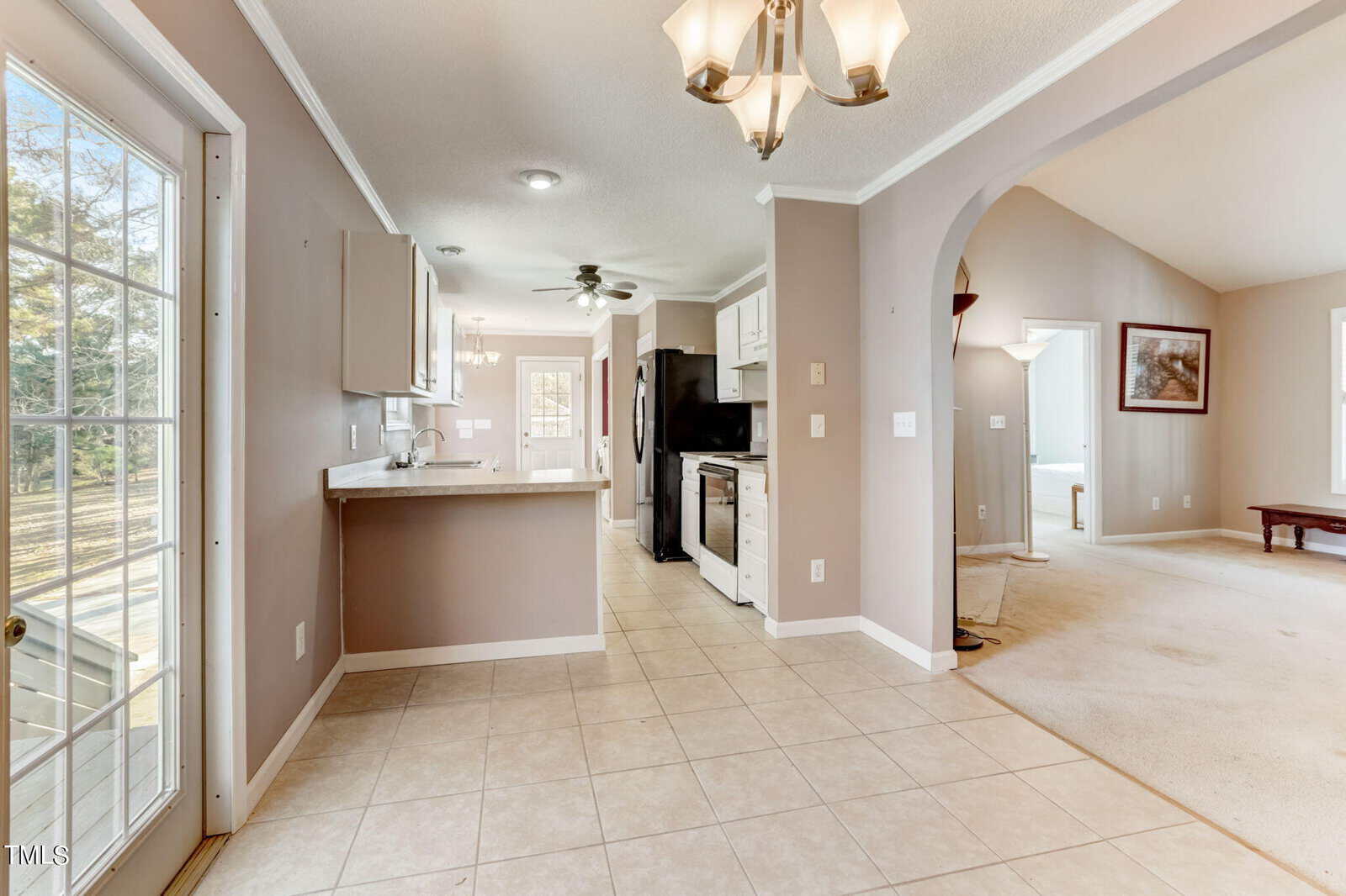 Spacious kitchen with tiled flooring, light brown walls, and white cabinets. A stainless steel refrigerator and stove are visible. A chandelier hangs above, and daylight streams in through a glass door and windows. An adjoining room is carpeted.