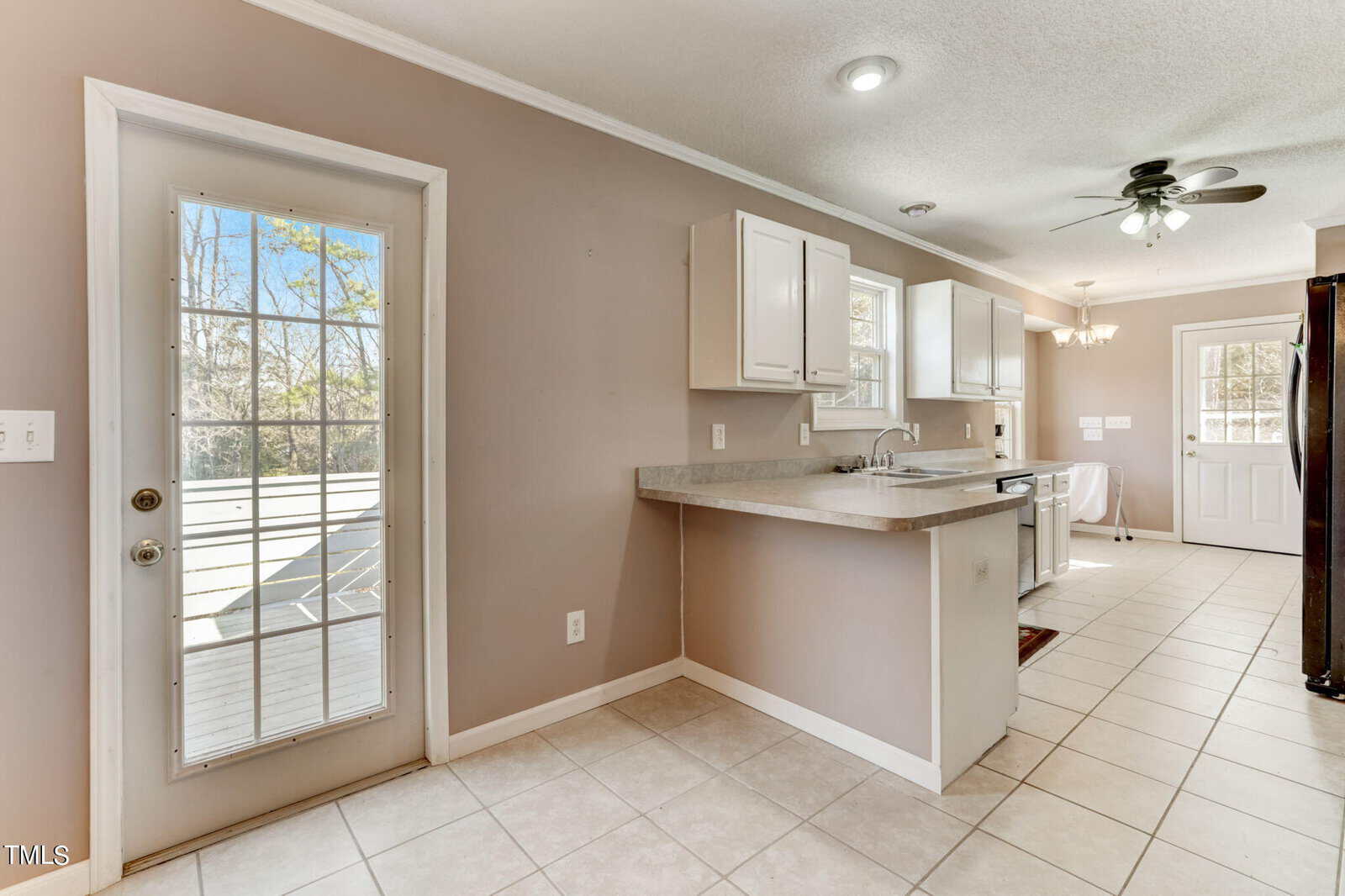 A bright kitchen with tan walls features white cabinets, a ceiling fan, and a countertop with a built-in sink. A glass door leads to a wooden deck outside. Natural light illuminates the space through multiple windows.
