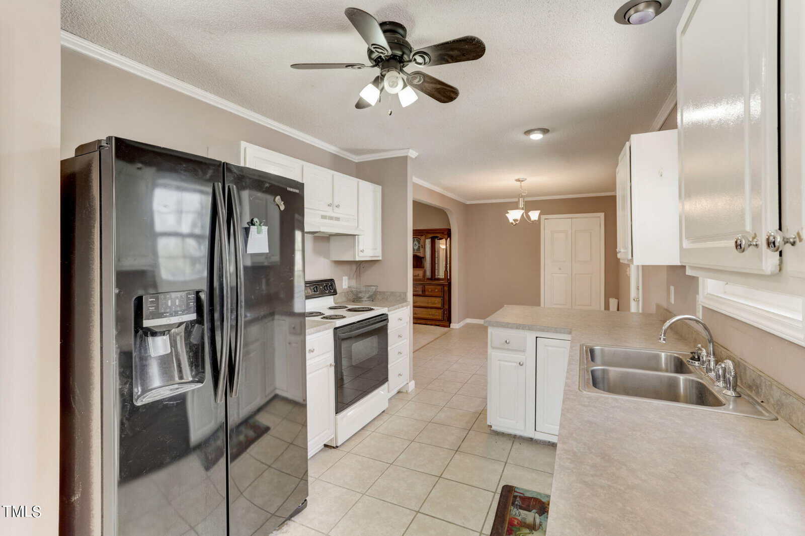 A modern kitchen with white cabinets, black fridge, and stove. A ceiling fan with lights hangs above beige tiled flooring. The kitchen has a double sink and ample counter space, leading to a doorway in the background.